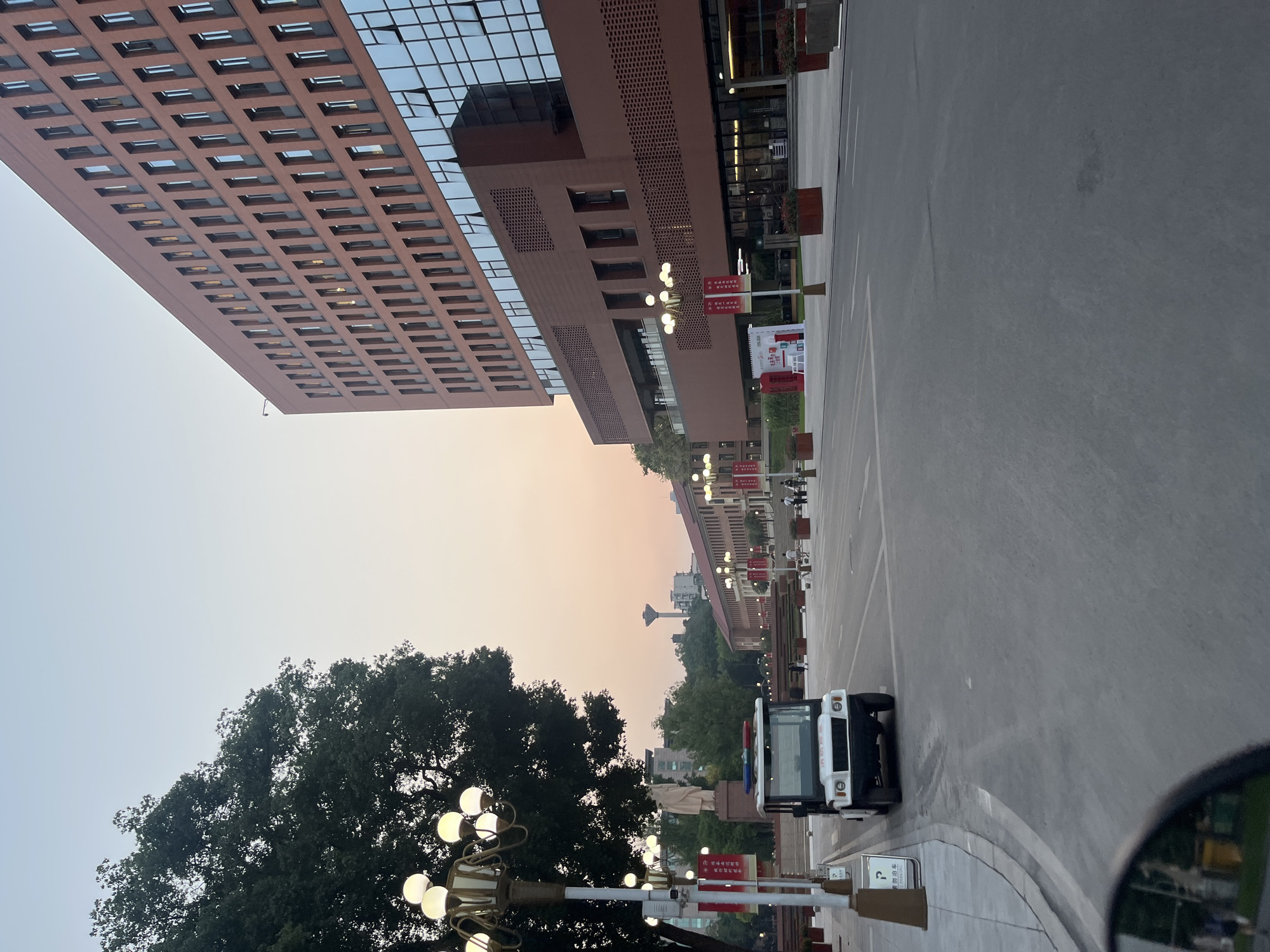This detailed image captures an urban or campus environment in Yuzhong District, China, during a transitional lighting period, likely dawn or dusk, evidenced by the clear sky's gradient from pale blue overhead to a soft orange-pink hue near the horizon.
The prominent feature on the right is a large, multi-story building with a reddish-brown facade. Its upper sections feature rows of rectangular windows, many reflecting ambient light. The lower two levels incorporate extensive glass panels and sections of intricate, ventilated brickwork or lattice patterns. Red banners with white Chinese text are visible at ground level near its entrance.
To the left, a mature, dense tree with dark green foliage occupies the upper left quadrant, partially obscuring the sky. In the foreground on the left, an ornate lamppost with five spherical white light fixtures stands prominently. Next to it, a white utility vehicle resembling a golf cart or security vehicle is positioned on the edge of the paved road. This vehicle features a blue and red light bar on its roof and visible Chinese characters on its front bumper. A "P" parking sign in Chinese is located on the curb near the vehicle. The very bottom-left corner of the image shows a partial reflection in a car's side mirror, capturing a distorted view of the immediate surroundings.
A wide, paved road or plaza extends from the foreground into the middle distance. Along this thoroughfare, additional identical ornate lampposts illuminate the path. In the mid-ground, to the left of the road, a large, light-colored statue of a standing human figure is situated on an elevated base, visible beyond the primary lamppost and tree. Further down the road, on both sides, other buildings with similar architectural styles are visible, and more red banners are displayed. In the far distance, silhouetted against the horizon, a tall, slender water tower with a spherical top is discernible. Several indistinct figures of people are sparsely distributed on the sidewalks and plaza areas in the mid-ground. Planters containing red flowers line various sections of the road and building fronts.