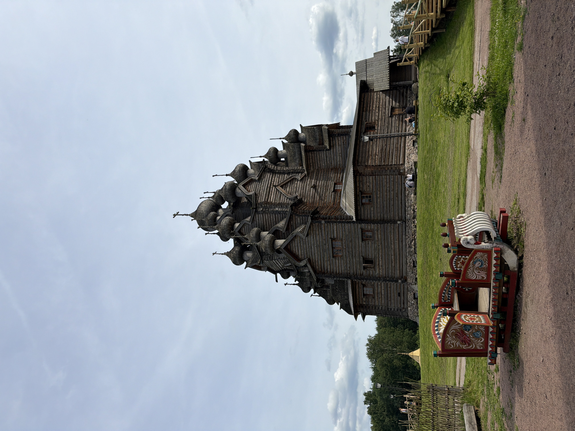 In the city of Imeni Sverdlova, Russia, a large, multi-domed wooden church stands on a grassy hill under a partially cloudy sky. In the foreground, a vibrantly painted wooden sled, possibly a decorative element or a prop, rests on a dirt path. The sled is adorned with red, white, and blue floral patterns and features a white, swan-like head at its front. Behind the sled, a low wooden fence encloses a grassy area. Further back, several people are visible sitting on benches near the base of the church, and others are ascending a wooden staircase leading to the church's entrance. Trees form a line in the background. The church itself is constructed from dark logs and features numerous small windows and intricately designed wooden elements.