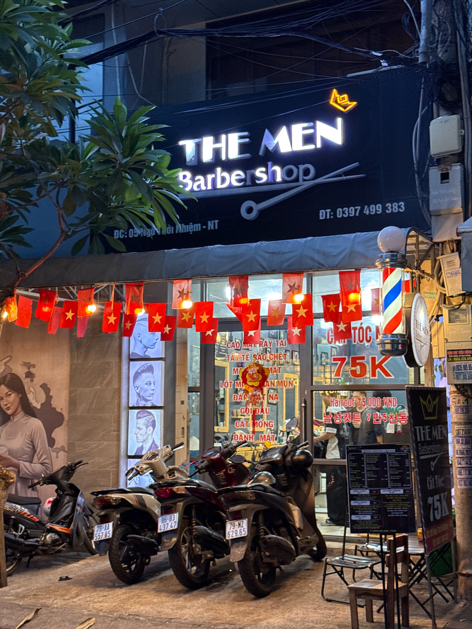 The image depicts the exterior of a barbershop named "THE MEN Barbershop" on a street in Nha Trang, Vietnam. The barbershop has a prominent black facade with white and light blue neon signage. A barber pole with red, white, and blue stripes is mounted vertically outside the entrance. Small red flags with yellow stars, characteristic of Vietnam, are strung across the front of the establishment, illuminated by small bulbs.
Several motorcycles are parked in front of the barbershop. The shop's windows display artwork and posters, including portraits of men with stylized haircuts and a price list for services. A menu board is visible inside, showing prices in Vietnamese Dong (VND) and Korean text. The street appears to be paved, and the overall lighting suggests it is either dusk or nighttime. The environment is urban, with visible power lines and electrical boxes on poles.