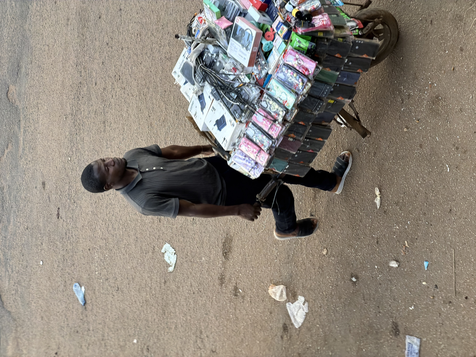 A lone Black male vendor pushes a two-wheeled cart laden with a variety of mobile phone accessories, including numerous phone cases and chargers, down a dusty, unpaved street in Bukuru, Nigeria. He is wearing a dark grey polo shirt and black pants, with dark sandals on his feet. The cart is positioned to his right as he walks away from the viewer and towards the right side of the frame. The street is littered with various pieces of debris, including plastic bags and wrappers. The overall scene suggests a typical urban street market environment in Nigeria.