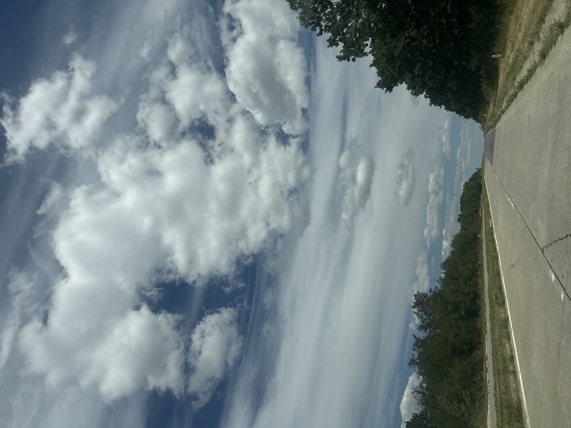 The image shows a two-lane road with a white line dividing the lanes and a white line along the right edge. The road is paved and appears to be cracked in places. On either side of the road, there are grassy verges and dense green trees. The sky is filled with a mix of fluffy white cumulus clouds and thinner, wispy cirrus clouds, against a blue sky. The scene is captured from a vehicle traveling on the road, looking forward. The location is identified as Sagaidacul Nou, Moldova.