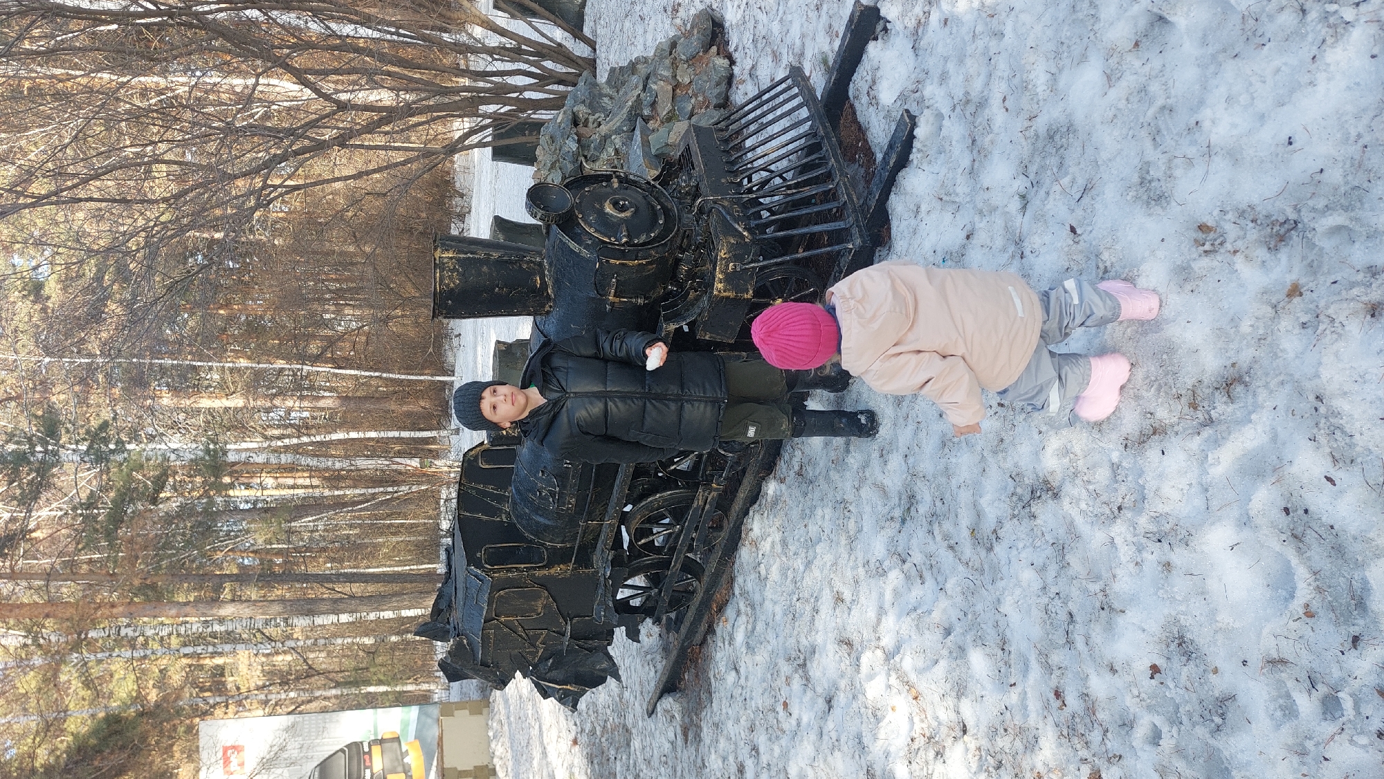 The image shows two children outdoors in a snowy environment. A boy wearing a black puffer jacket and dark hat is standing next to a black, antique-style train locomotive display. He is holding a ball of snow. Beside him is a younger child wearing a light pink jacket, grey pants, and pink boots, bending over in the snow. The background includes leafless trees, suggesting it might be winter or early spring. The location appears to be a park or public space with the locomotive as a display piece. Based on the style and appearance of the display and environment, it may be located in the city of Novosibirsk, Russia.