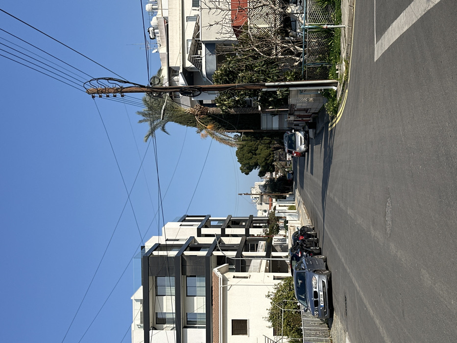 This image depicts a residential street scene under a clear blue sky, likely in Egkomi, Nicosia, Cyprus, as suggested by the location context. The photograph is taken during bright daylight, indicated by strong sunlight and distinct shadows cast on the asphalt road and buildings.
The street, which curves gently into the distance, is lined with a mix of multi-story buildings. On the left, a modern apartment block with white facades and prominent black-framed balconies and window units stands out. Below it, a traditional white-painted building is visible. On the right side of the street, more residential buildings of varying ages are present, some with external water tanks or solar panels on their roofs.
A wooden power pole, heavily laden with multiple overhead electrical wires, dominates the mid-ground on the right. Several palm trees and other deciduous trees, some appearing bare, are interspersed among the buildings and along the street, suggesting a Mediterranean climate.
Parked cars are visible along both sides of the road. On the left, a dark blue BMW sedan with a visible license plate "WZF 330" is parked, along with a motorcycle behind it. Further down the street on the left, another silver car is parked. On the right side of the street, closer to the foreground, a silver hatchback is parked near the power pole. Another car is seen further down the street on the right.
The road features double yellow lines indicating no parking zones and white road markings in the foreground. No people are visible in the scene, which captures a quiet moment in a residential area. A small sign with partially visible text, possibly "SOL," is seen on the modern building on the left.