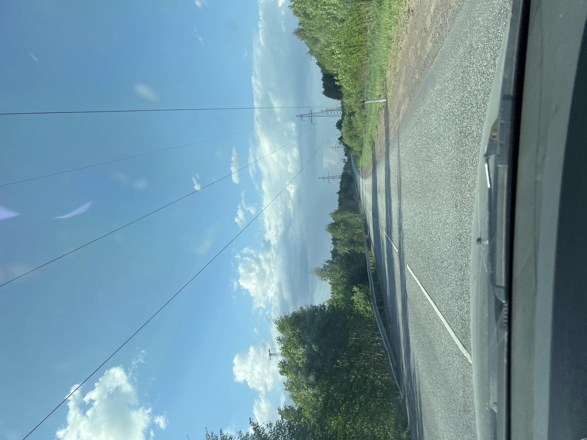 A low-angle, tilted shot taken from inside a vehicle shows an asphalt road with a white dividing line. Guardrails run parallel to the road. Power lines stretch across a partly cloudy, blue sky. Transmission towers are visible in the distance. The road is flanked by green vegetation, including trees and possibly crops. Based on the visible elements of foliage, road structure, and power lines, and referencing the city location of Chaikovsky, Russia, it's likely the shot shows a roadway in the surrounding area, possibly a rural or semi-rural route on the outskirts of the city. There are no visible people.