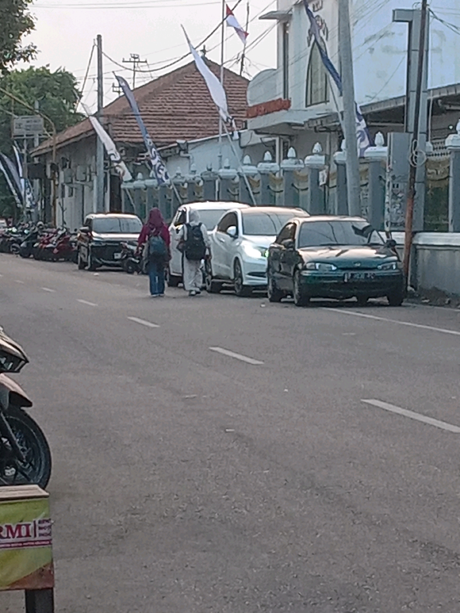 The image captures a street scene in Yogyakarta, Indonesia, likely during daylight. In the foreground, a portion of a motorcycle's front wheel and fender is visible on the left, next to a box with text that appears to be in Indonesian. The road, paved with asphalt, has white lane markings. Several parked cars are lined up on the right side of the road. Among them, a dark green sedan is prominent, with its front facing the camera. Behind it, a white sedan is also parked. Two individuals are walking on the road between the parked cars. One person, wearing a red headscarf and blue pants, carries a backpack. Another individual, wearing white clothing and a head covering, also has a backpack. The background reveals buildings with tiled roofs and a facade that suggests a public or religious structure, possibly a mosque, adorned with flags. Trees are visible on the far left. Power lines and poles are also present, indicating urban infrastructure. The overall impression is a mundane street view with typical urban elements.