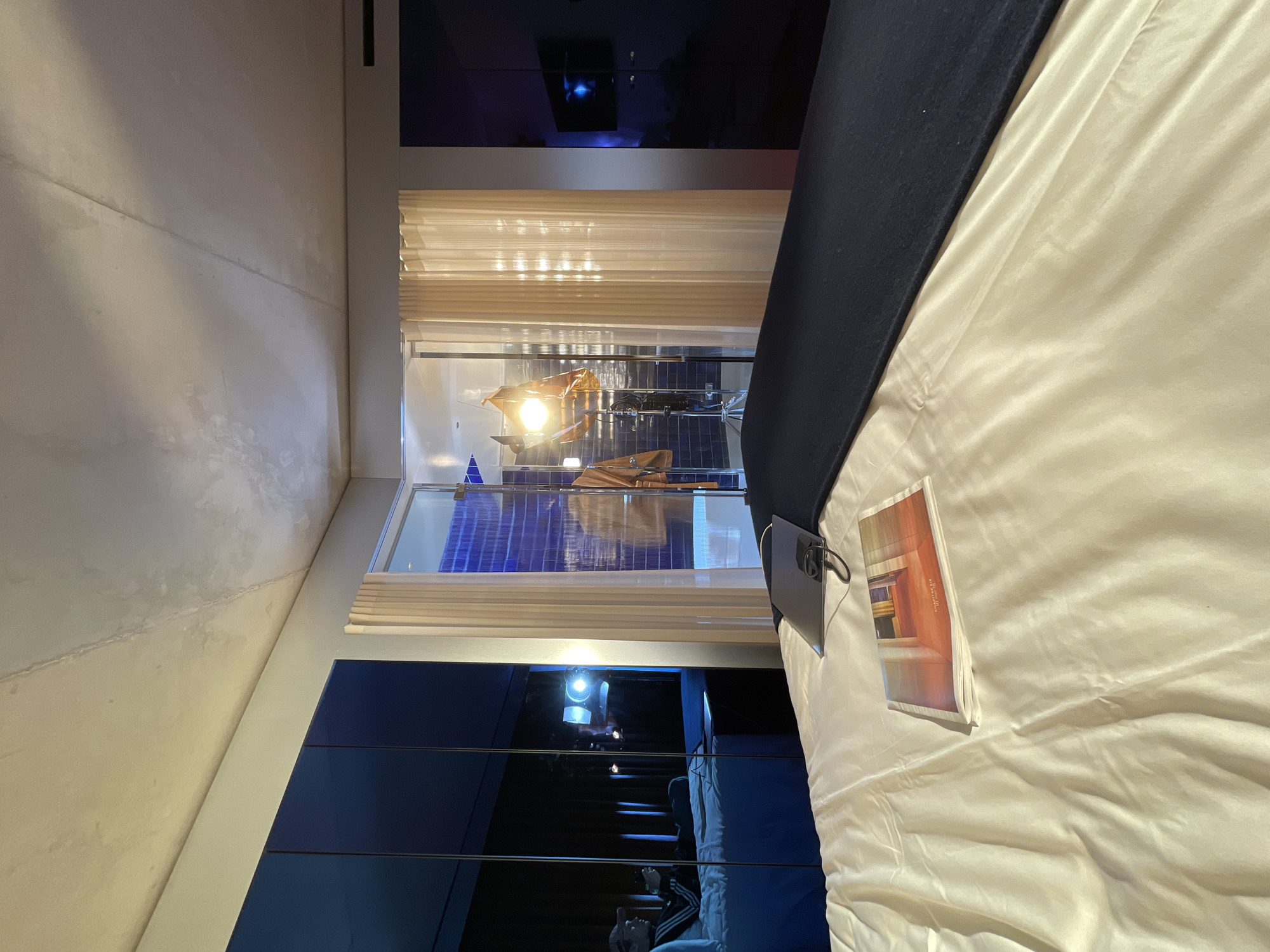 A bedroom interior is shown, presenting a view of the Bulgari Hotel in Paris, France. A bed with white sheets and a dark blue throw lies in the foreground, where a magazine and a tablet device rest on the bedding. Through a window equipped with beige blinds, a bright light shines. In the room below, blue tiles are visible, and a figure is seen on a staircase. Dark blue panels or furnishings are present on the walls, contrasting with the white ceiling and adjacent wall.