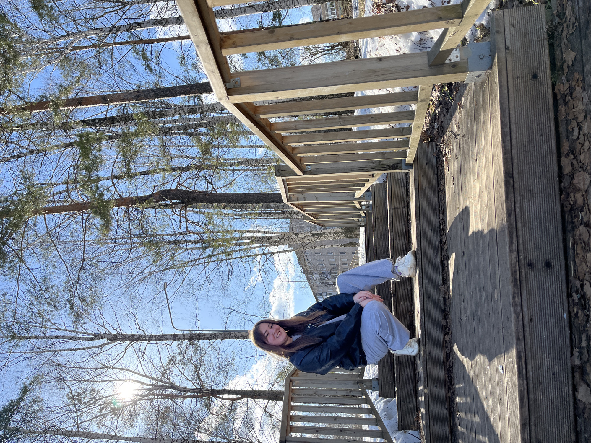 The image captures a young woman sitting on a wooden staircase outdoors in Chaikovsky, Russia. She is wearing a dark jacket, grey sweatpants, and white sneakers. The staircase is made of planks, and there is some snow visible on the sides. In the background, there are trees and a clear blue sky. The sun shines through the branches.