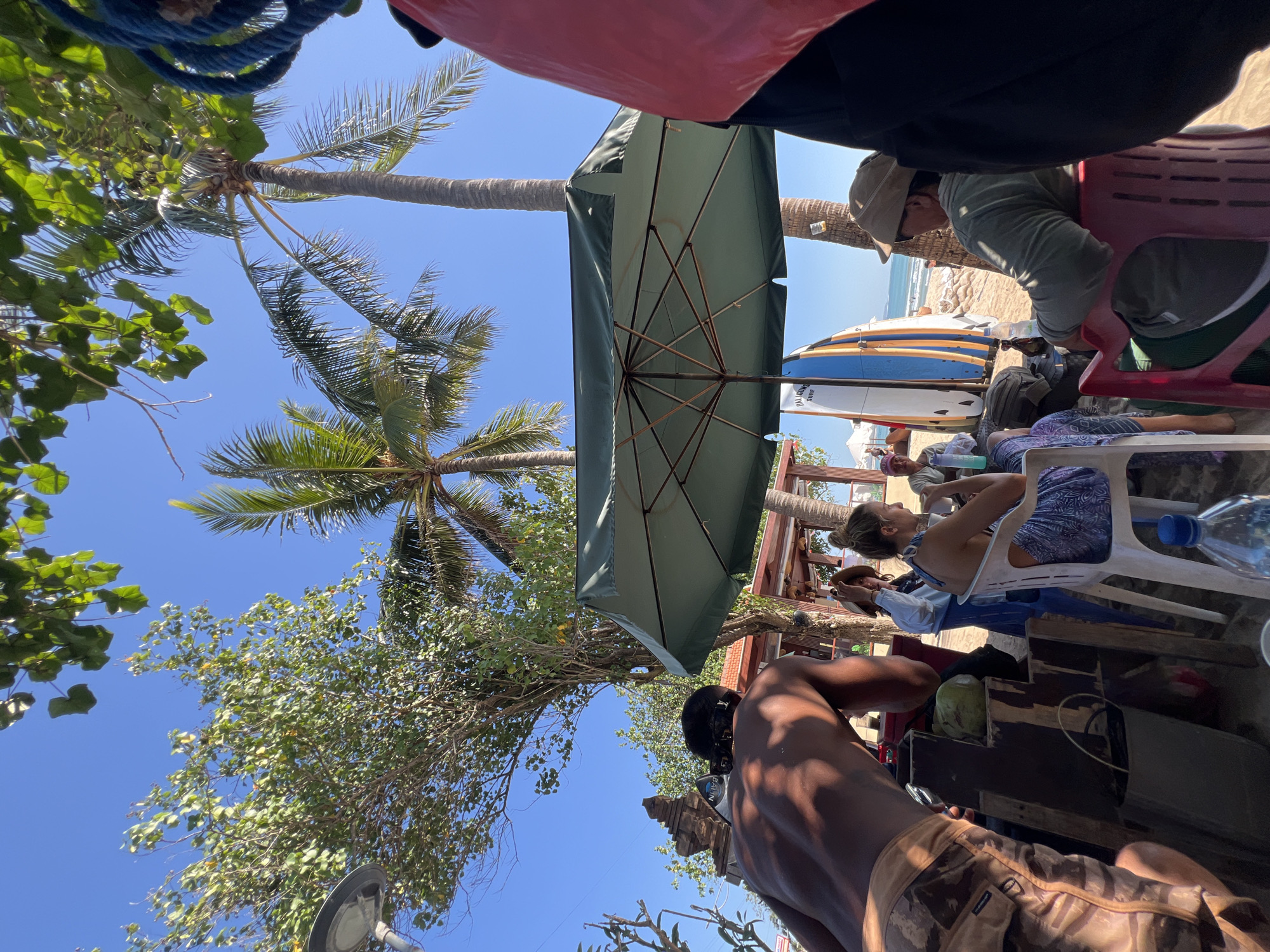 In Kuta, Indonesia, under a bright blue sky, a group of people is gathered on a beach, seeking shade from a large green umbrella. Two surfboards, one white with blue and orange stripes and another mostly blue, stand upright near the umbrella. In the foreground, a person with dark skin and short black hair pulled back, wearing camouflage-patterned shorts, is partially visible, leaning forward. Nearby, a whole coconut sits on a wooden surface.
To the right, under the umbrella, a woman with blonde hair tied up, wearing a blue and white patterned dress, sips from a white cup. She is seated on a white plastic chair. Beside her, a man wearing a light-colored baseball cap and a green long-sleeved shirt is seated on a red plastic chair. Behind them, a tropical temple-like structure with ornate carvings is visible through the trees.
The environment suggests a beachside setting, with sand visible and the ocean in the background. Palm trees and leafy green trees provide additional shade. The overall scene depicts a relaxed gathering at the beach.