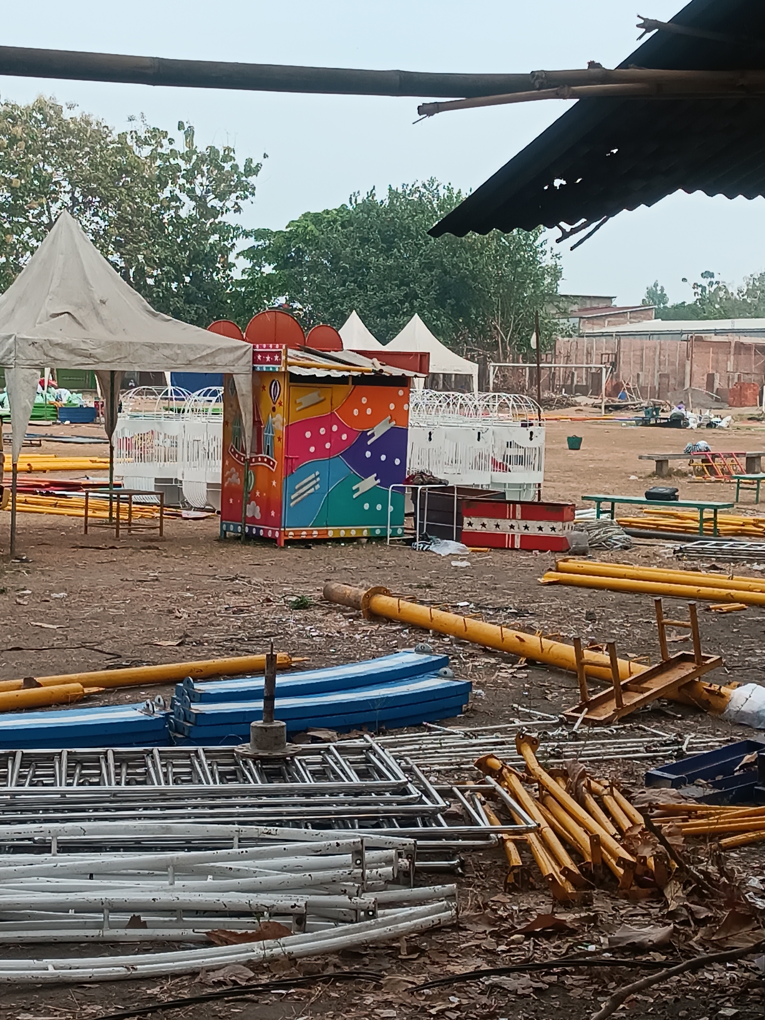 This is a wide shot of an outdoor area with temporary structures and amusement ride components. In the foreground, there are stacks of disassembled metal framing, some painted yellow and others white. There are also blue and white rectangular platforms. Behind these, a colorful, small amusement ride structure with a patterned exterior is visible. To its left, a white tent-like structure is set up. Further in the background, more white, open-frame amusement ride structures are present. The ground is covered in dirt and fallen leaves. In the distance, buildings and trees are visible. The scene suggests a temporary setup or dismantling of an amusement area in Pleret, Indonesia.