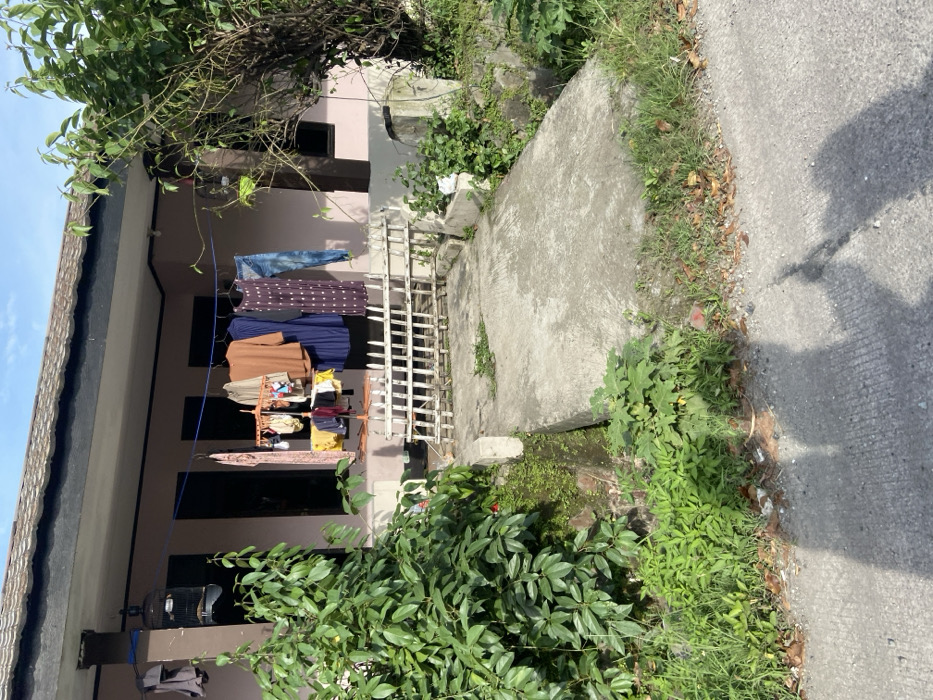 The image shows a concrete walkway leading to a house in Jatiluhur, Indonesia (latitude: -6.578, longitude: 107.436). The walkway is made of concrete and has a slight incline. The house is a single-story building with a brown tiled roof. There is a clothesline strung across the front porch, with various articles of clothing hanging from it, including a blue shirt, a purple dress, and a pair of jeans. The house is painted a pale pink color. The walkway is lined with a white picket fence. There is a birdcage hanging on the wall of the house. There is also a small green bush growing on the left side of the walkway. The walkway ends at a concrete pad in front of the house. In front of the house, there are a variety of plants and shrubs. There is a large tree to the right of the house. The tree is blocking part of the house from view. There is a shadow of a person on the ground near the edge of the walkway. The scene is quiet and peaceful, with no people visible.