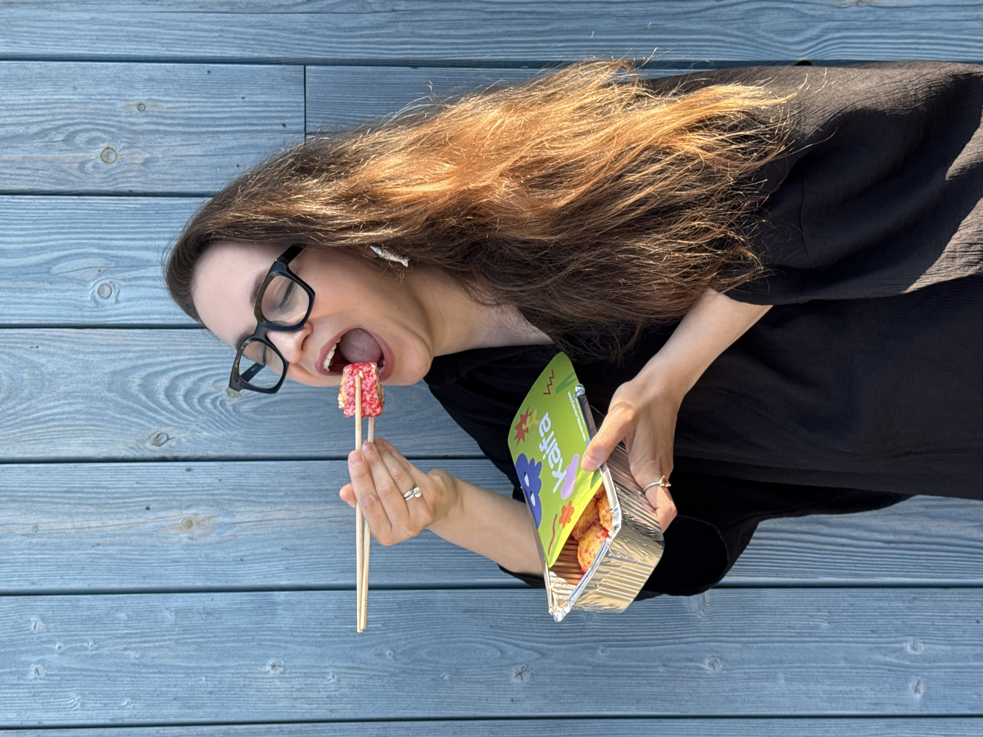 In Chaikovsky, Russia, a woman with long, brown hair and glasses is about to eat a piece of sushi. She holds the sushi with chopsticks and has her mouth open. She is wearing a black top and rings on her fingers. The sushi is in a foil container. The background is a blue wooden wall.
