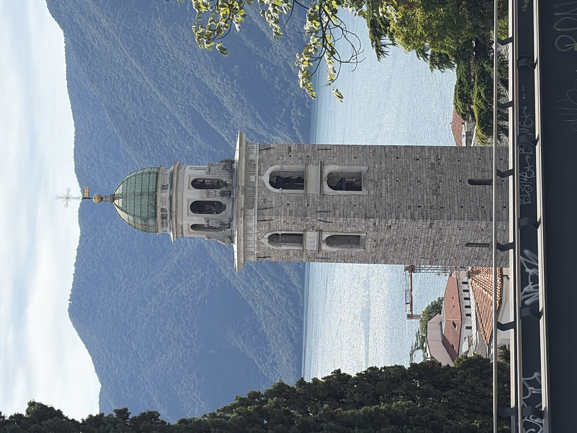 The image shows a stone bell tower with a green dome, topped with a cross, situated in the city of Lugano, Switzerland. The tower is made of grey stone blocks and features arched windows, some of which contain bells. The bell tower is positioned in the foreground, with a body of water, likely Lake Lugano, sparkling in the background. Behind the lake, lush green mountains rise, their slopes covered in dense vegetation. In the middle ground, red-tiled roofs of buildings are visible, along with a construction crane bearing the name "MEDIC". To the left of the tower, a tall, dark green coniferous tree stands prominently. The scene is bathed in natural daylight under a partially cloudy sky, suggesting it was taken during the day. A dark metal railing with graffiti is visible in the bottom foreground.