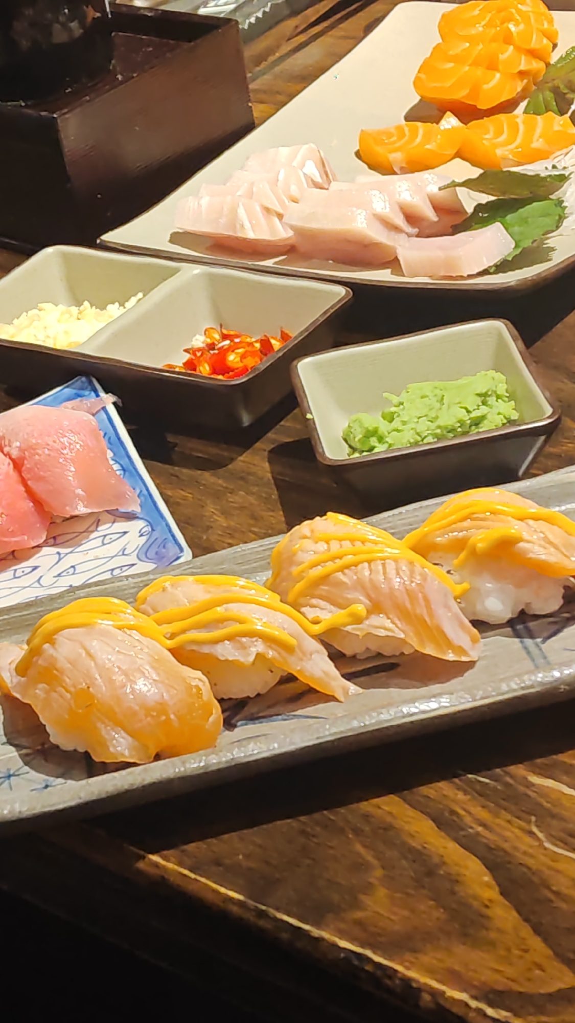This image depicts a close-up, high-angle view of a sushi platter and various accompaniments, likely in a restaurant setting. The foreground features several pieces of nigiri sushi, each topped with a generous drizzle of yellow sauce, possibly mayonnaise or a cheese sauce. Behind the nigiri, a small blue and white patterned dish holds three slices of raw pink fish, likely tuna. Further back, two square, dark-rimmed dishes contain finely minced garlic and chopped red chili peppers, respectively. To the right of these, another dark-rimmed dish holds a vibrant green mound of wasabi. Dominating the background is a long, white rectangular plate holding slices of raw salmon, arranged in a fanned pattern with a few green garnishes. The food is laid out on a wooden surface, suggesting a table. The lighting is warm and somewhat diffused, creating a rich, appetizing glow on the food. The scene does not provide any explicit indicators of its specific location within Đà Nẵng, Vietnam; however, the style of the sushi and accompaniments is consistent with common offerings in Japanese restaurants found in urban centers like Đà Nẵng.