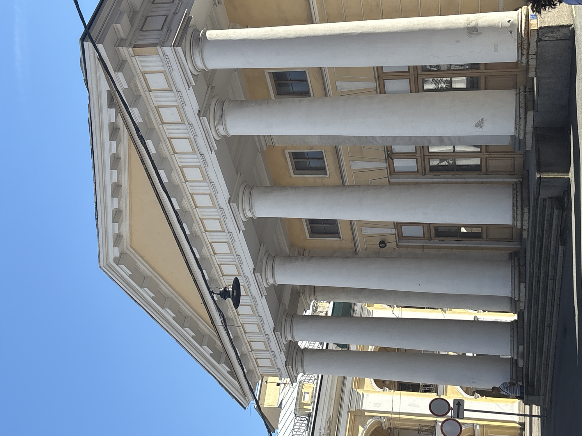 The image shows the south facade of the Russian National Library, a neoclassical building in Saint Petersburg, Russia. The facade features a prominent portico with six large, fluted Ionic columns supporting a triangular pediment. The building is painted a pale yellow, with white decorative elements. Several windows and doors with wooden frames are visible on the ground level. In front of the building, a set of stone steps leads up to the entrance. To the left of the steps, a man in a striped shirt stands near a street sign with two circular signs and an arrow. The sky is clear and blue.