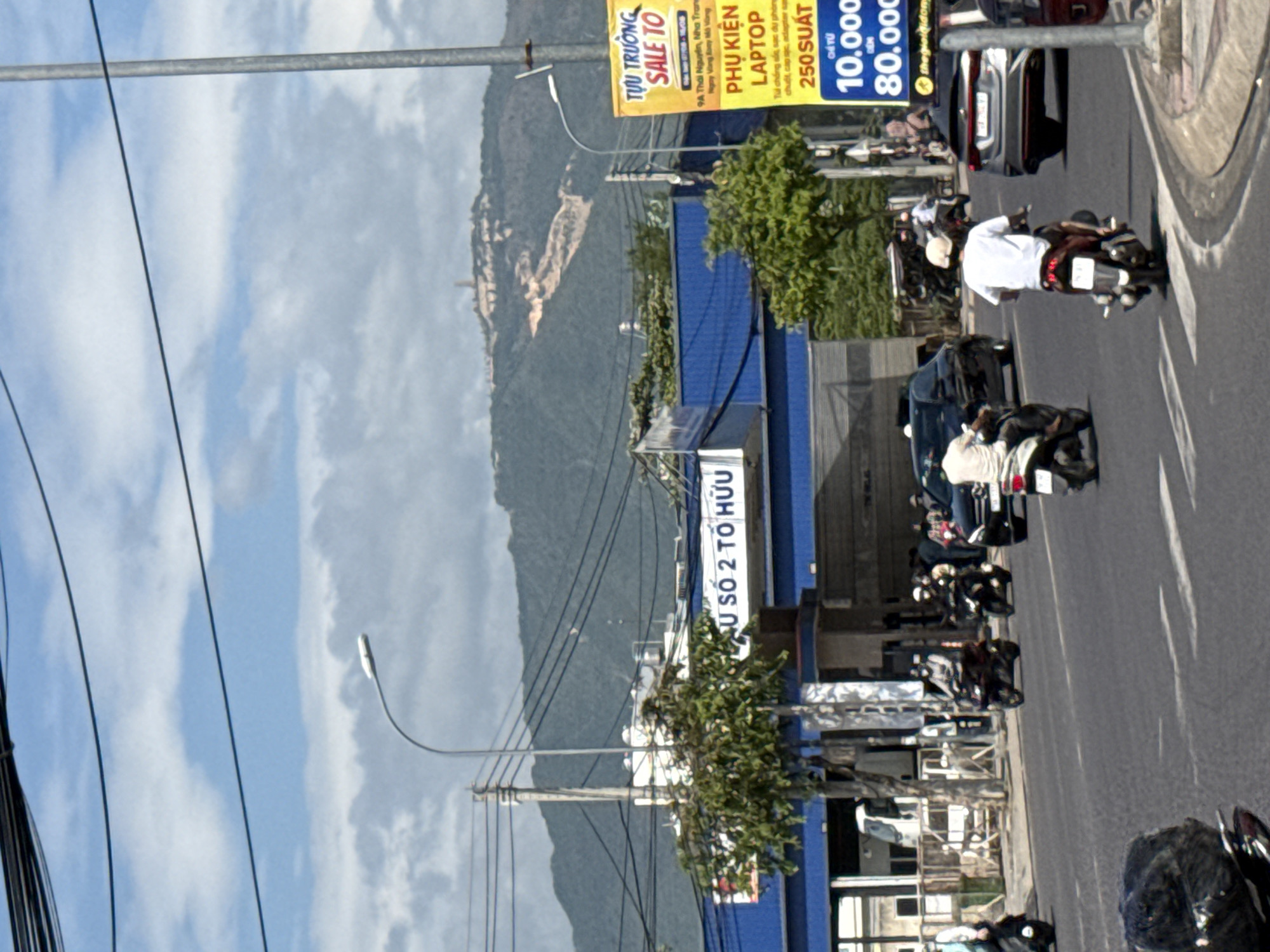 This image captures a street scene in Nha Trang, Vietnam, featuring a busy road with vehicles and pedestrians, likely in the vicinity of a commercial area given the presence of signage and a building with blue awnings.
In the foreground, a dark-colored motorcycle with a person on it is partially visible on the left. Moving further down the road, a person on a white motorcycle is seen from behind, wearing a white shirt and a cap. To the right of this motorcycle, a silver sedan is driving, and further to the right, a portion of a red car is visible.
On the road, several other motorcycles are present. One black motorcycle with a rider wearing a white shirt and helmet is moving away from the viewer. Another motorcycle, a dark color with a rider, is in the middle lane. In the background, there are more vehicles, including a white truck with a large cargo area, parked or moving slowly.
A building with a blue facade and white lettering that reads "SỐ 2 TỔ HỮU" is visible, suggesting it's a business or service establishment. There is also a large yellow advertisement sign on the right side of the image, displaying text in Vietnamese, including "SALE TO" and "PHỤ KIỆN LAPTOP" (Laptop Accessories), along with prices in Vietnamese Dong (e.g., "250.000", "10.000", "80.000").
The environment includes a paved road with lane markings, sidewalks, trees, electrical poles with numerous wires overhead, and a distant view of a lush green mountain range under a partially cloudy sky. The overall scene depicts an active urban street environment.