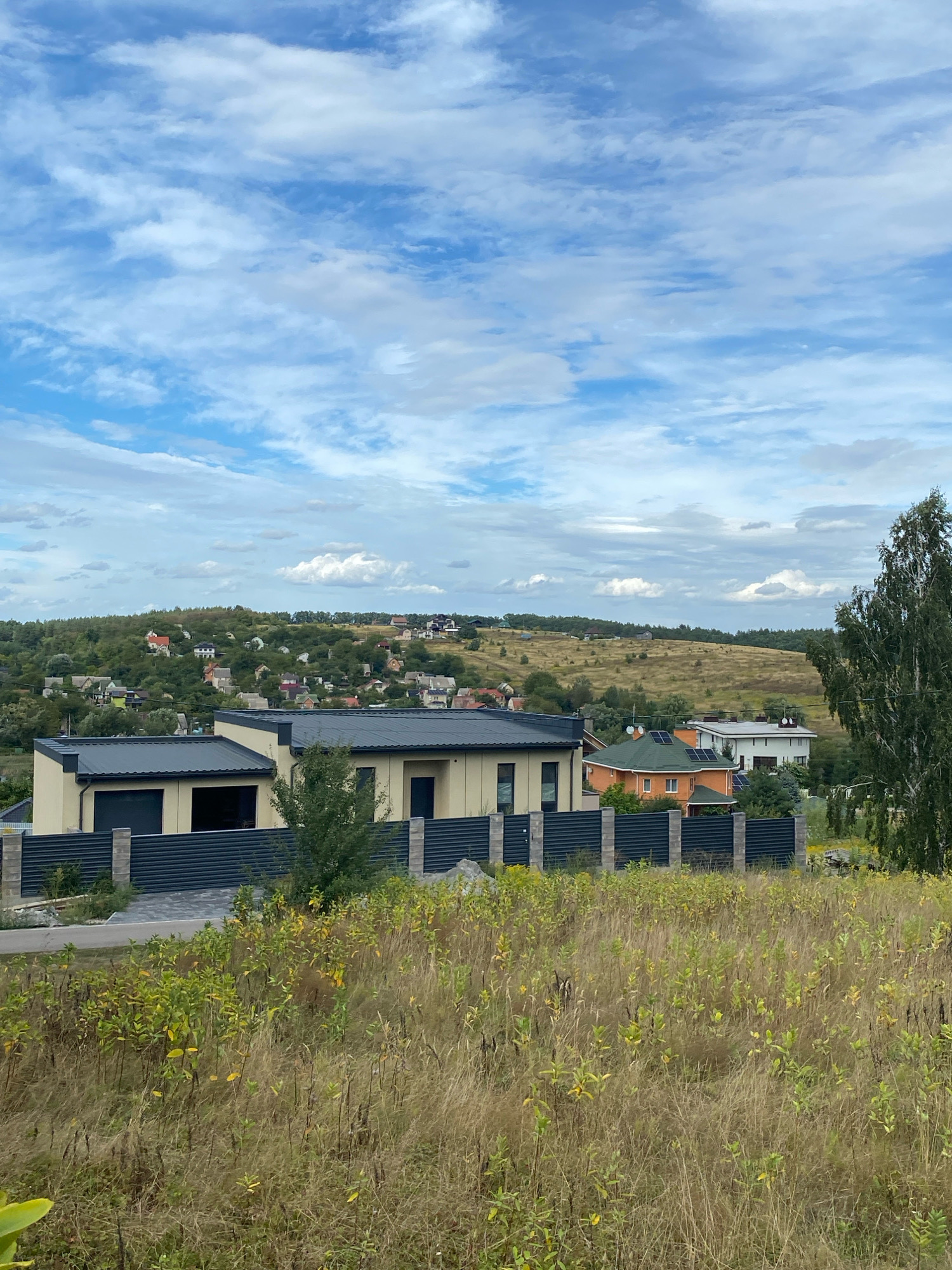 The image depicts a rural landscape in Horbovychi, Ukraine, under a partly cloudy sky. In the foreground, tall, dry grass and scattered green and yellow foliage dominate the scene, suggesting a late summer or early autumn setting. A dark metal fence runs horizontally across the lower portion of the image, separating the foreground from a property beyond.
Behind the fence, a modern, light-colored house with a dark roof and large windows is visible. Adjacent to this house is a vibrant orange house with a green roof, equipped with solar panels. Further in the background, a cluster of houses and buildings are nestled among rolling, tree-covered hills, forming a small village or community. The hills in the distance are a mix of green foliage and dry, golden grasses. A tall, slender tree with light-colored bark stands to the right of the frame, its branches reaching towards the sky. The overall impression is a tranquil, suburban or semi-rural environment. There are no people or animals visible in the image.
