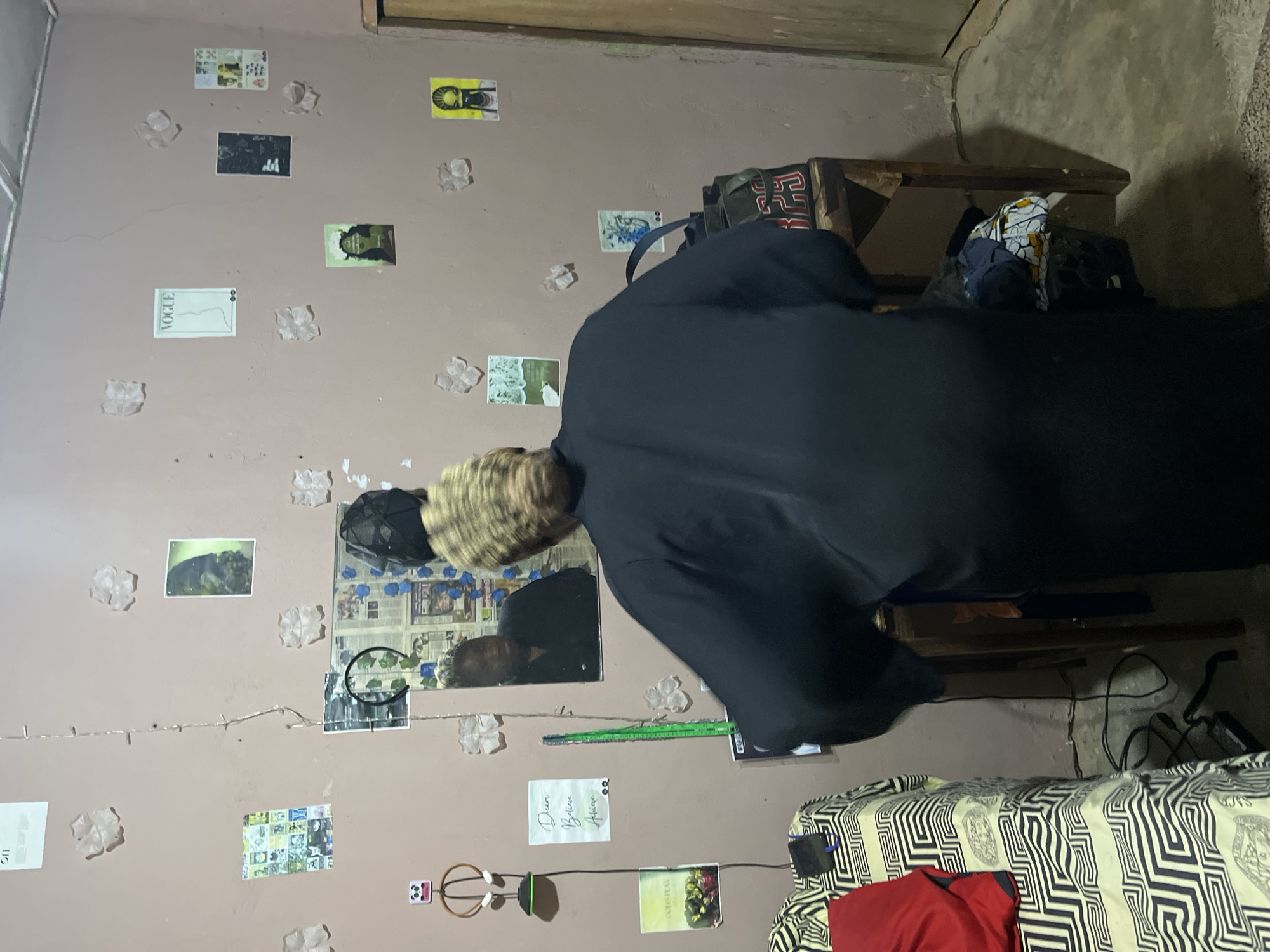 A woman wearing a black robe is standing in front of a mirror in a sparsely decorated room. The room appears to be in a residential setting in Rafin Par, Nigeria. The walls are painted a light pink and are adorned with various posters and what appear to be decorative flower cutouts. In the mirror, the woman is visible from the chest up, and she appears to be looking into the mirror. To the left of the mirror, there is a bed with a patterned sheet and a red garment on it. A desk is positioned to the right of the mirror, with a bag and some other items on it. The floor is concrete, and there are power cords visible on the floor. The lighting in the room is dim, with a string of fairy lights also present on the wall.
