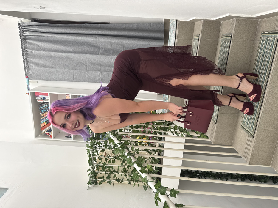A woman with purple and pink hair, wearing a sheer burgundy dress and matching platform sandals, poses on a staircase. She holds a burgundy handbag. The background features a curtained window, a bookshelf, and a decorative wall covered in faux ivy. The staircase has white railings and patterned tiles on the steps. The lighting is bright and evenly distributed.