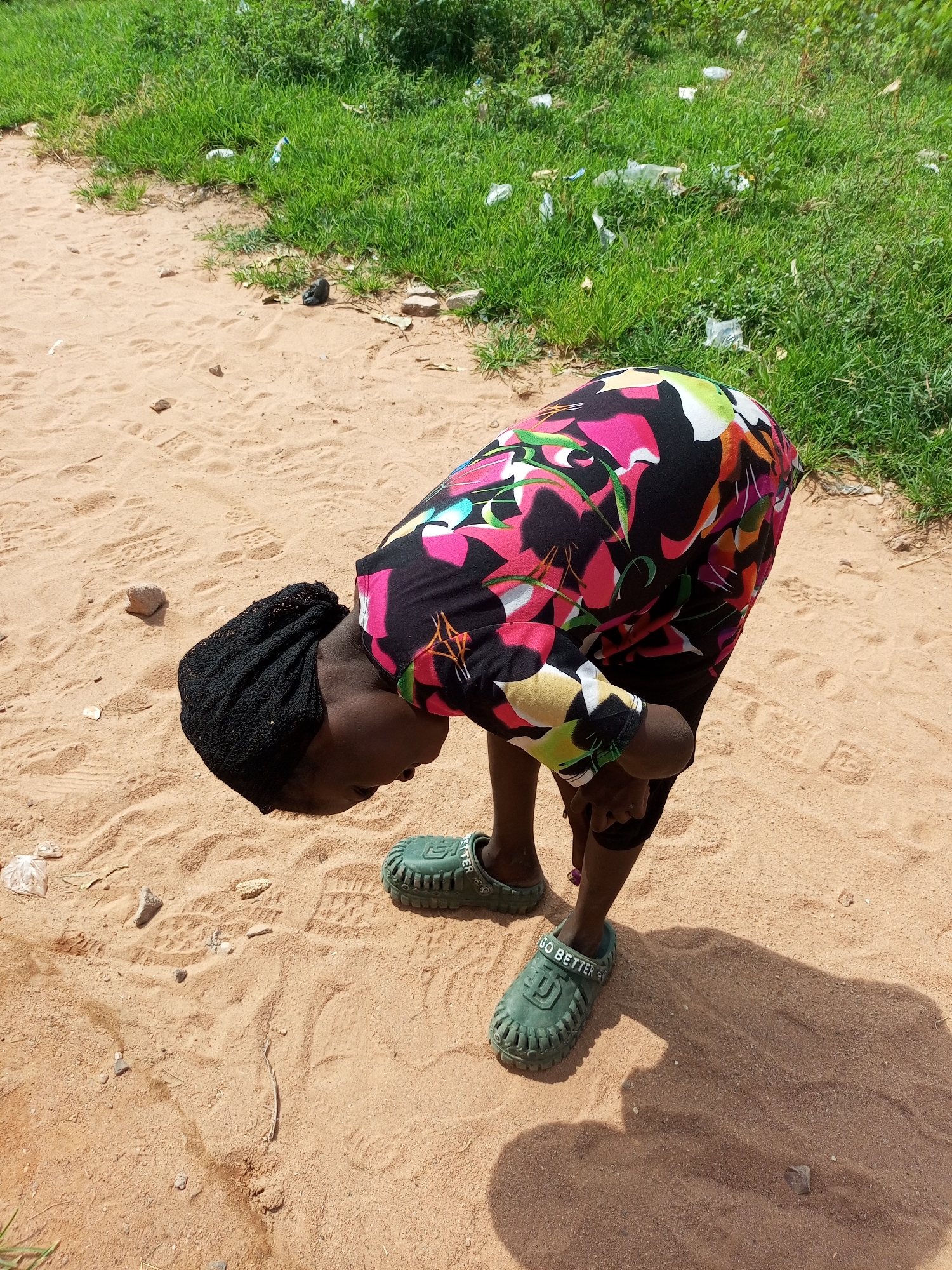 A young African person, wearing a black headscarf and a colorful, floral-patterned dress, bends over in a sandy area. The person's feet are clad in green clogs that read "GO BETTER". The ground is covered in fine sand with visible footprints and small stones. To the right and in the background, a patch of vibrant green grass grows, with some scattered white plastic waste visible amongst the blades. The scene suggests an outdoor, possibly rural or semi-urban, setting in Tempure, Nigeria, characterized by its sandy terrain and natural vegetation.