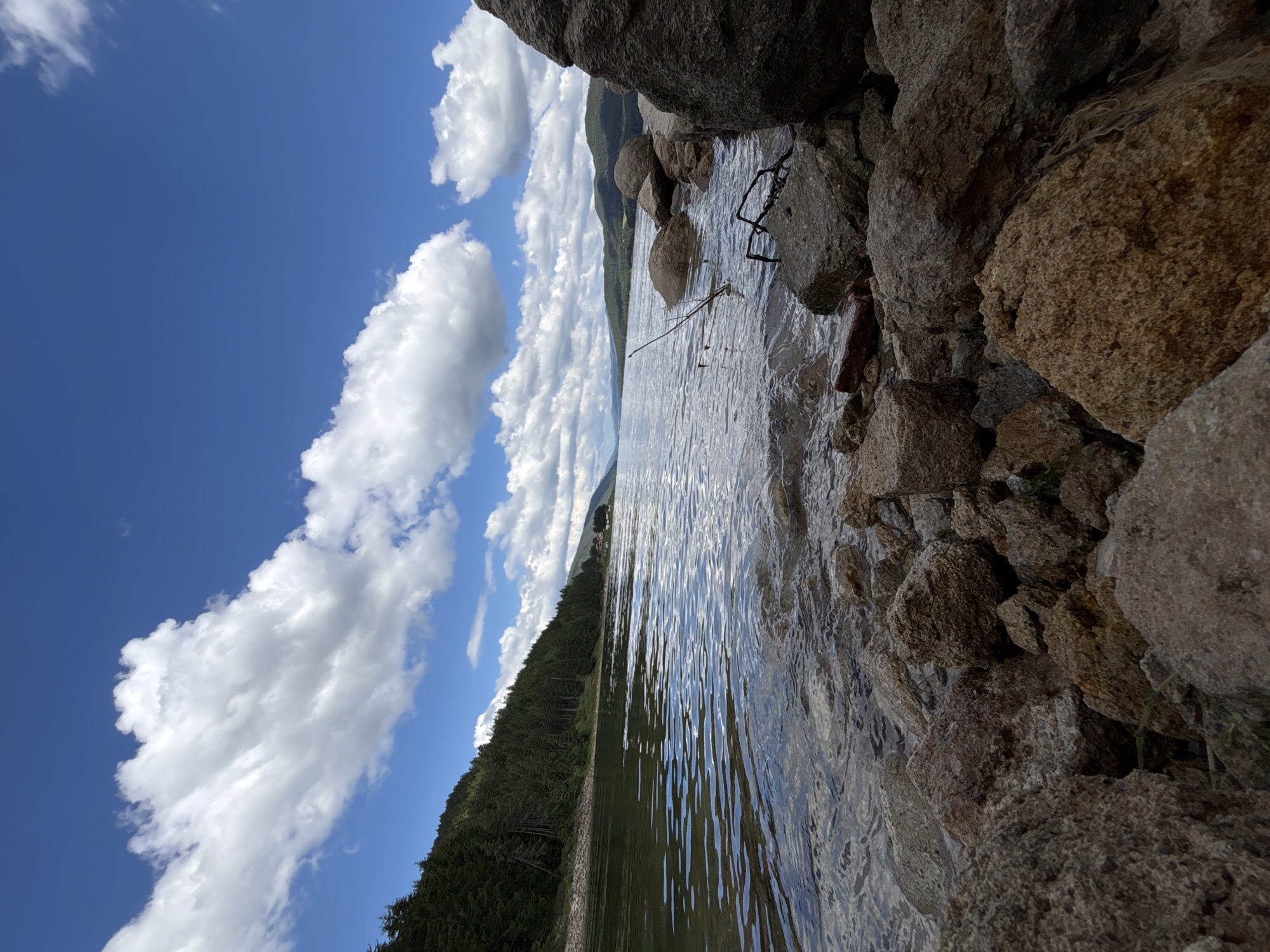 An eye-level, wide-angle shot captures a scenic view of a calm lake bordered by a dense, evergreen forest on the left and rolling, tree-covered hills in the distance. The foreground is dominated by a rocky shoreline, with various sizes of weathered stones and a few scattered twigs partially submerged in the shallow, clear water. The lake's surface is gently rippled, reflecting the bright blue sky dotted with fluffy white clouds. A few distant buildings are visible nestled amongst the trees on the far shoreline. The overall impression is one of natural tranquility under a partly cloudy sky.