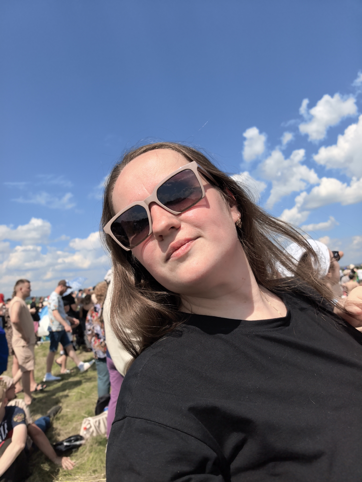 A woman wearing sunglasses and a black t-shirt is pictured in a selfie at a sunny outdoor event. The background shows a crowd of people and a blue sky with clouds. The location is identified as the city of Nestiukovo, Russia, suggesting a community gathering or festival. The woman is the primary subject, with the crowd and environment providing context.
