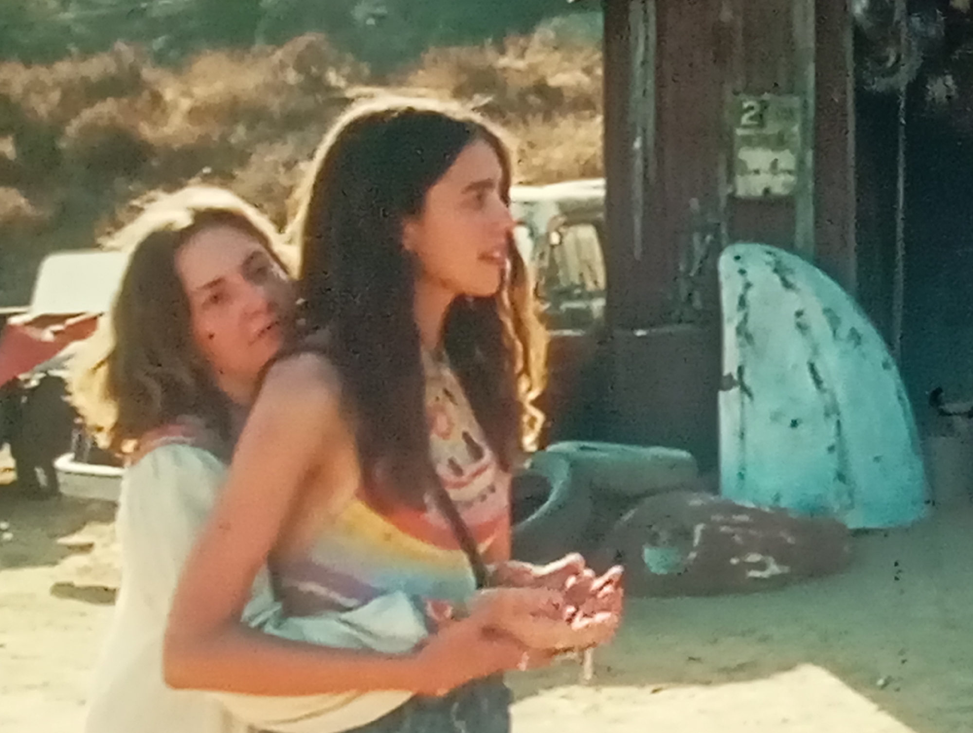 Two young women are embracing outdoors. The woman in the foreground, on the right, has long dark hair and is wearing a rainbow-striped tank top. She is looking to her right, with her hands clasped in front of her. The woman behind her has lighter brown hair and is wearing a white top. She is looking to her left. They appear to be in a rural or outdoor setting, with a building and some objects in the background, including what looks like tires and a white, amorphous object. The lighting suggests it is daytime, likely late afternoon due to the warm tones. The image quality is grainy, typical of older photographic film. The scene is devoid of any specific indicators pointing to the city of Sofiivska Borshchahivka, Ukraine, or any urban environment.
