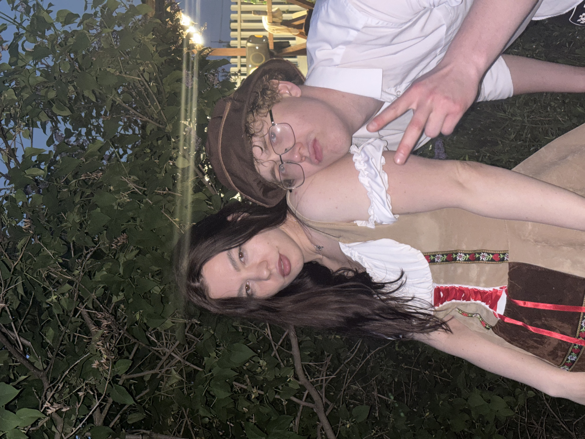 Two individuals are visible in the image. A male wearing a brown hat and glasses is leaning his head on the shoulder of a female. The female has long, dark hair. Both are wearing what appears to be traditional clothing, possibly related to Oktoberfest. They are positioned in an outdoor environment with a leafy tree visible in the background. The photograph seems to have been taken in Пальники, Russia. The man's hand is positioned, making a V shape.
