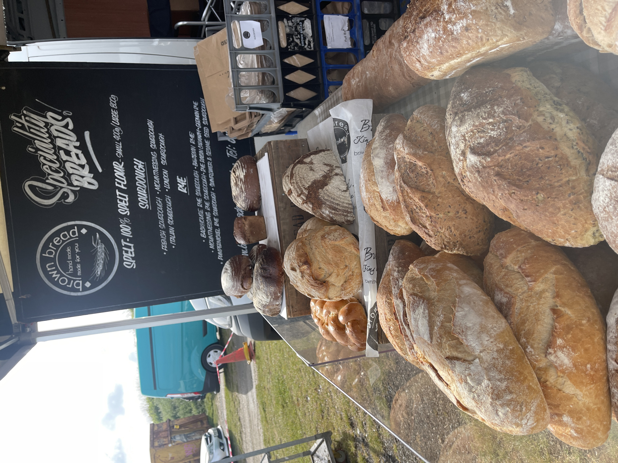 The image shows a bread stall at a market, likely in Sevenoaks, United Kingdom. The stall features a variety of specialty breads, including sourdough and rye varieties. A sign advertises "Speciality Breads" from "brown bread," emphasizing "hand made, made for you". The breads are neatly displayed on a transparent surface. The sign details the types of bread available, including sourdough options like "French Sourdough" and rye options like "Bavarian Rye". The breads appear to be made with 100% spelt flour. In the background, there are bread racks and containers. Outside the market stall, there's a blue vehicle and surrounding greenery, suggesting an outdoor market location.