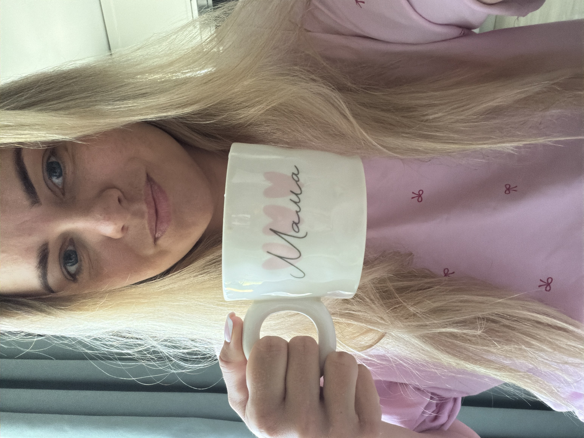 A blonde woman with blue eyes is holding a white mug with the word "Mama" written on it in Russian with pink heart accents. She is wearing a pink top with a bow pattern. The background appears to be an interior shot with a neutral wall and a possible window or curtain. The image provides no specific information that can locate it in the city of Votkinsk, Russia.