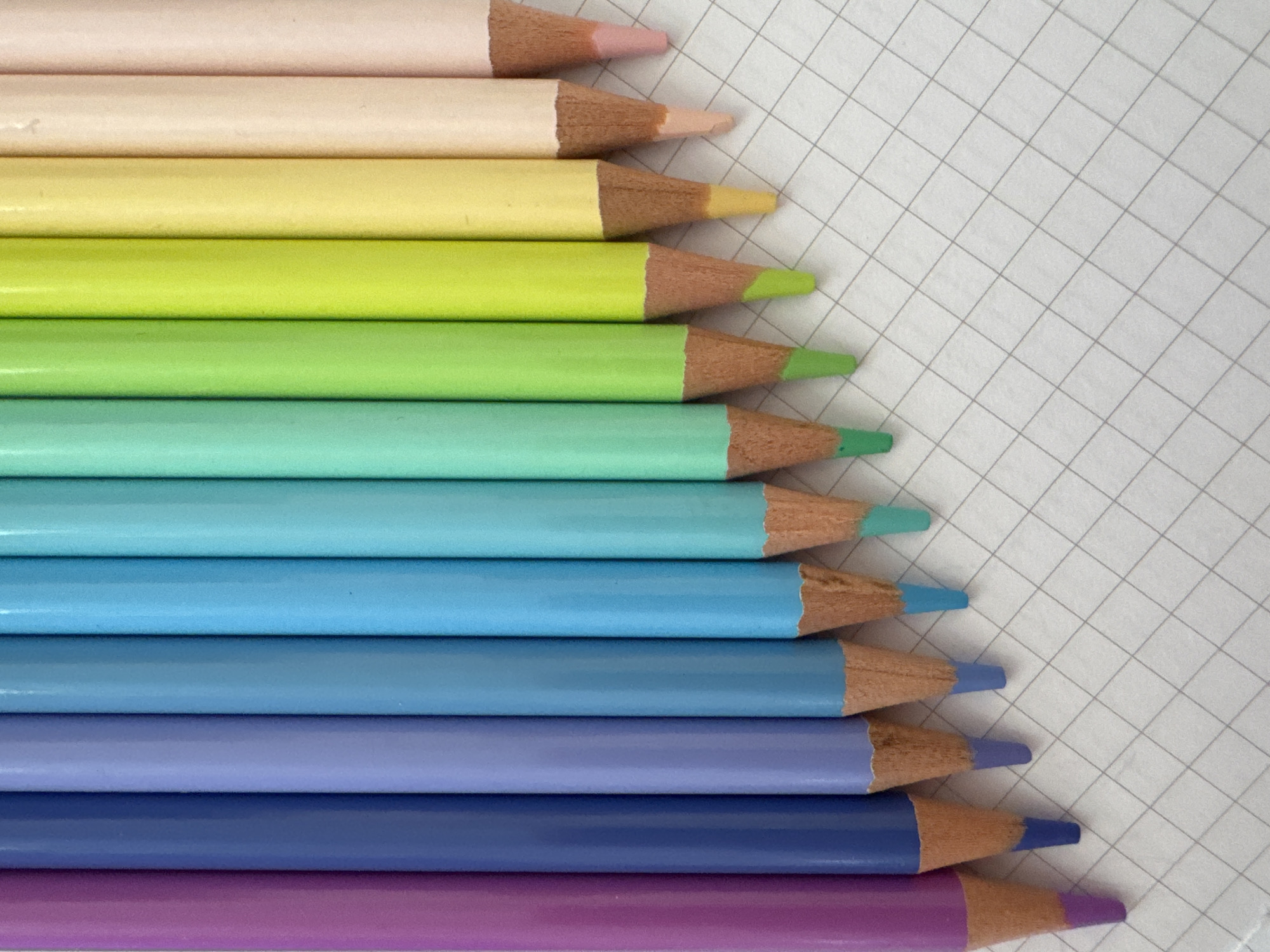 The image displays a collection of colored pencils arranged diagonally on a white, gridded surface, possibly notebook paper. The pencils are laid out in a row, with their sharpened tips pointing towards the lower right corner of the frame. They are ordered in a gradient of colors, starting with light pink at the top and transitioning through various shades of yellow, green, and blue, ending with purple at the bottom.
The pencils are of similar size and shape, with a visible wooden casing and a colored tip. The surface beneath them shows faint gray grid lines. The lighting appears to be even and diffused, suggesting an indoor setting or an overcast day. There are no people or animals present in the image, and no specific event is depicted. The arrangement of the pencils suggests a still life composition, focusing on the colors and textures of the art supplies. No text is visible in the image.