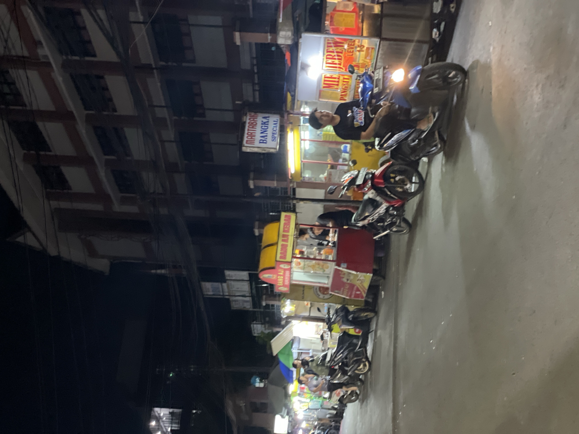 Captured at night in Central Jakarta, Indonesia, the scene presents a bustling street filled with food vendors and parked motorcycles. Illuminated signage indicates the presence of food stalls, with one labeled "MARTABAK BANGKA SPECIAL" and another "MIE JBREW LEVEL PANGSIT." A man is seated on one of the motorcycles. Further down the street, food vendors operate from their carts, and there are numerous motorcycles parked alongside. The urban environment is dark, with light emanating from street lamps and the food stalls themselves. The architecture consists of multi-story buildings along the side of the street.