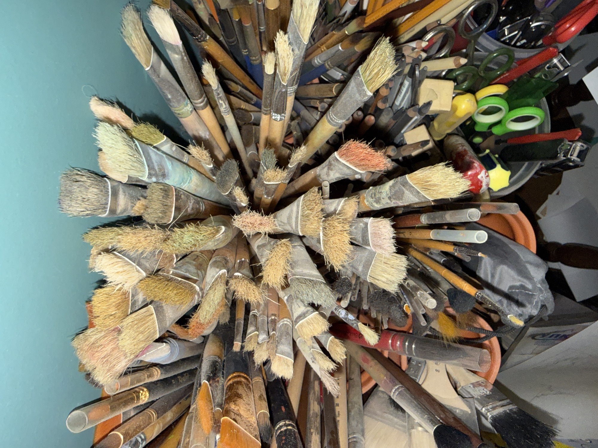The image depicts a tightly packed collection of paintbrushes of various sizes and types, viewed from above, fanned out radially from the center. The brushes have wooden handles, some with visible paint or wear, and ferrules connecting them to bristles that vary in color from natural to reddish-brown. Many of the brushes are dusty or stained, indicating use. Mixed in with the brushes are other art supplies, including pencils, scissors, and possibly some small tubes or containers. The overall impression is one of a well-used artist's studio or workspace. The background appears to be a teal-colored wall. The image does not provide any information about the location in Votkinsk, Russia.