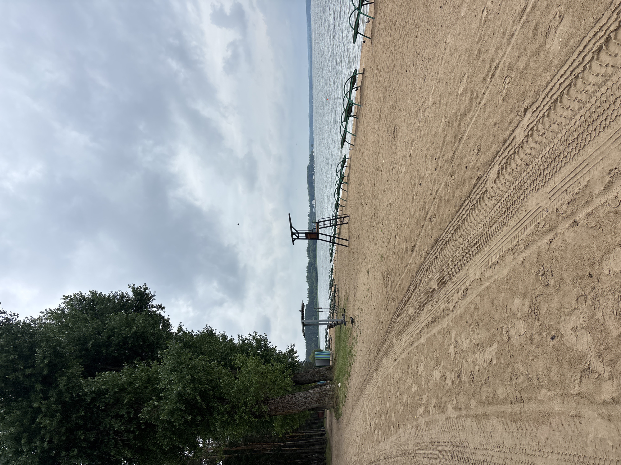 The image depicts a sandy beach area with a large body of water in the background, under a cloudy sky. The beach is marked by tire tracks from vehicles in the sand. To the left, several large trees line the edge of the beach. In the middle ground, there are rows of empty green lounge chairs set up on the sand. A lifeguard tower with a red railing stands on the right side of the beach. The water appears calm, and distant land with some structures is visible on the horizon. The overall atmosphere is overcast and quiet, suggesting an unpopulated or off-season beach. The location is described as Ждановіцкі сельскі Савет, Belarus, indicating a rural or suburban administrative area.