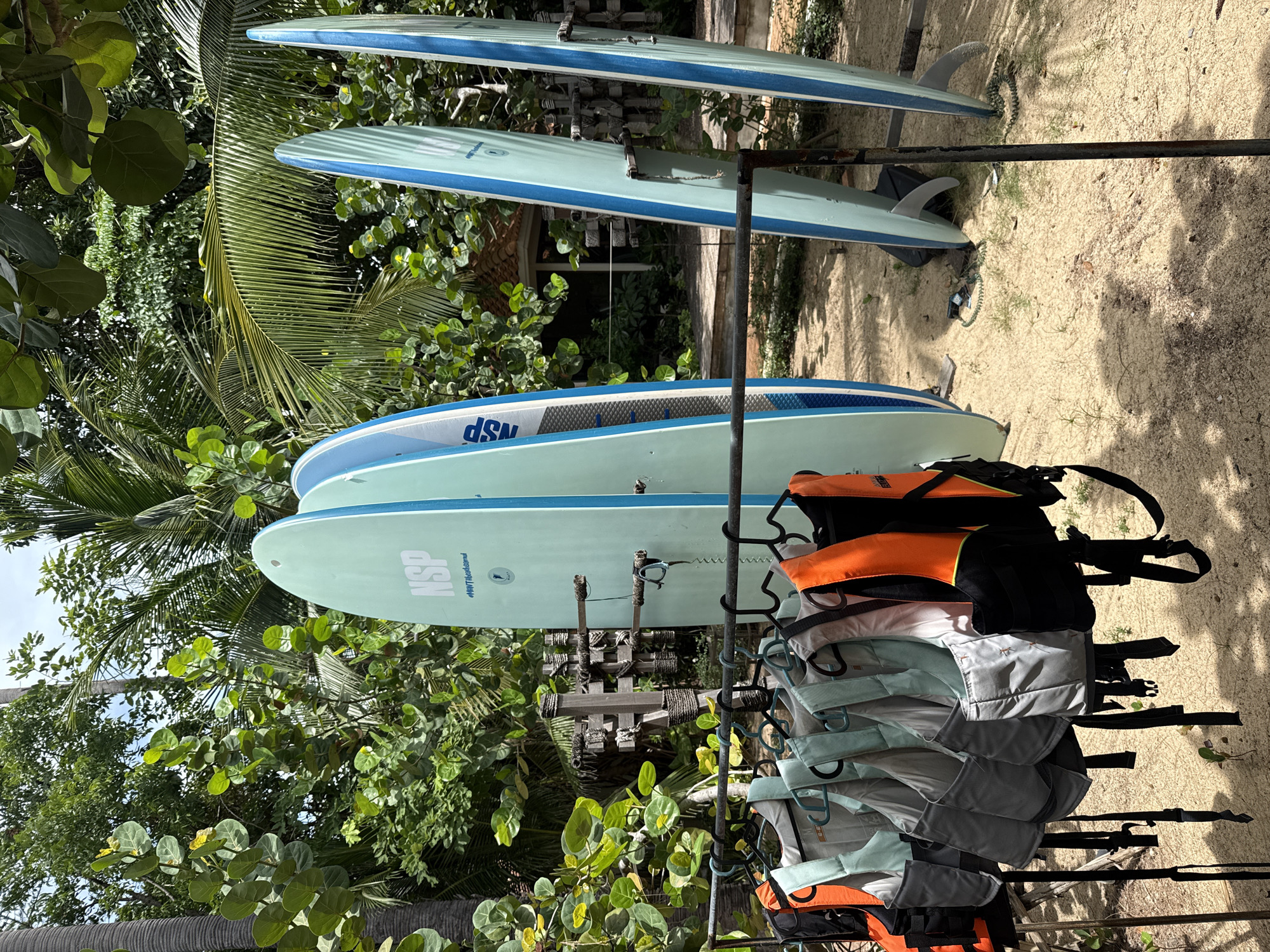 The image depicts an outdoor scene in a tropical environment, likely in the Ko Samui District of Thailand, with lush green foliage and palm trees. In the foreground, a rack displays several life vests in various colors, including orange and gray, hung on hangers. Behind the life vests, several paddleboards are standing upright. These paddleboards are predominantly light blue and white, with some featuring blue trim. The ground is covered in sand and scattered green grass. In the background, a wooden structure with a tiled roof is visible amidst the dense vegetation, suggesting a resort or a recreational area. The overall impression is one of a setup for water sports and outdoor activities in a warm, sunny climate.