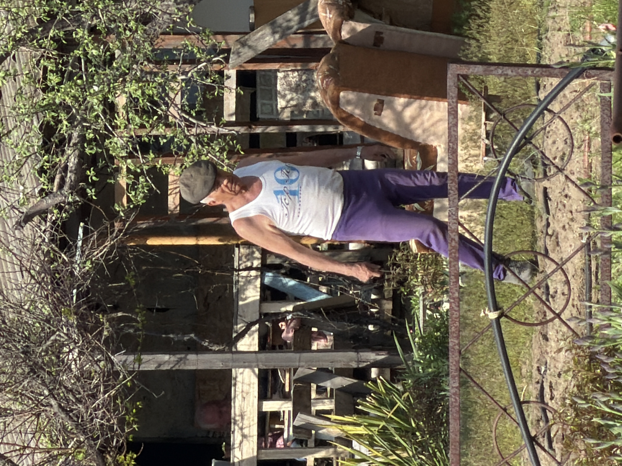 A middle-aged male wearing a gray cap, white tank top, and purple pants stands in an outdoor setting. He is facing left with his right arm extended. The background includes a dilapidated structure with wooden planks, furniture, and vegetation. In the foreground, a metal gate stands among grass and other plants. It is unknown if the location is Kamyshin, Russia.