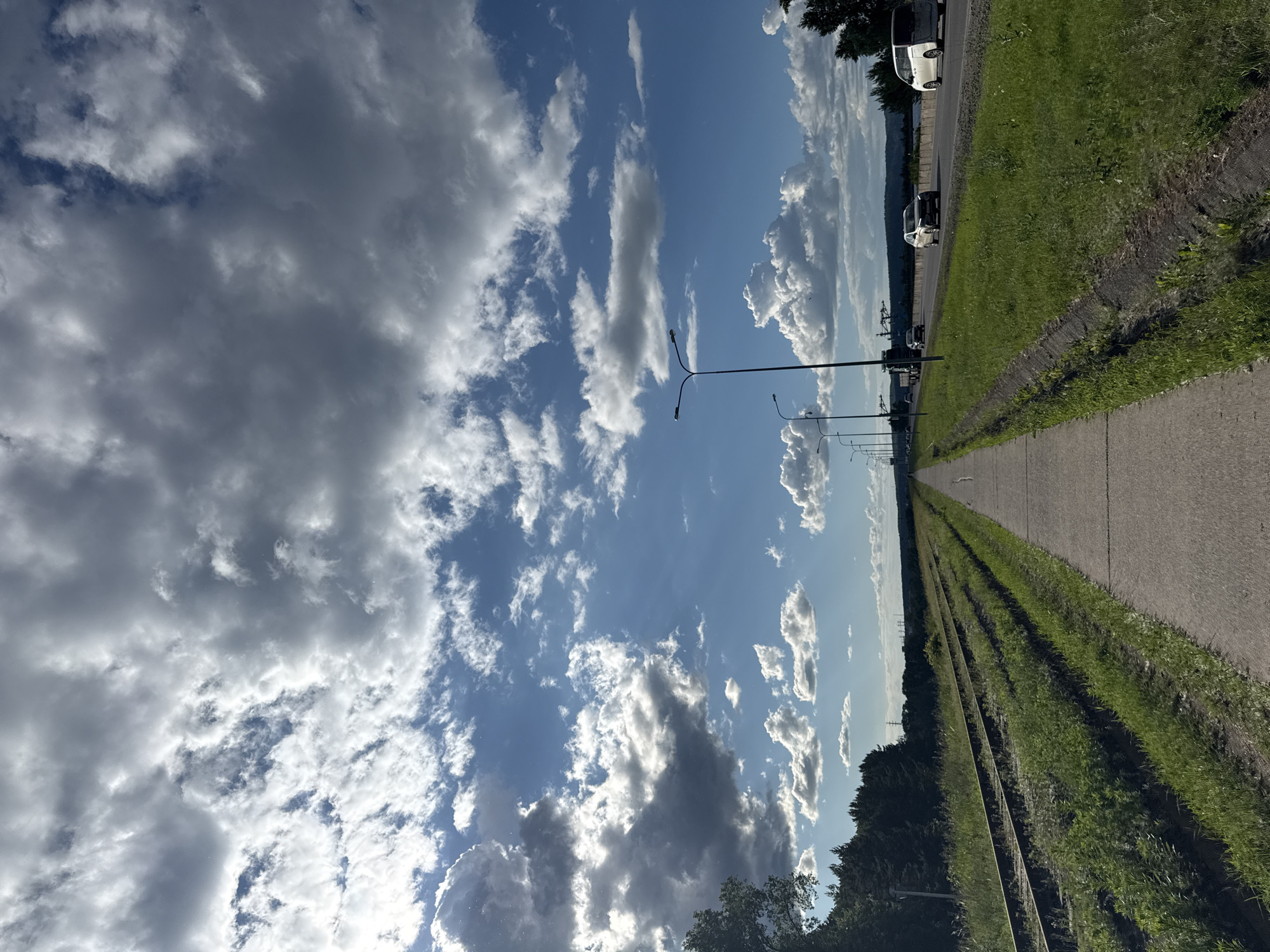 The image depicts a scenic view of a paved pedestrian path running parallel to a set of railroad tracks on the left and a highway on the right. The path leads into the distance, creating a strong sense of perspective. The sky is filled with dramatic clouds, with patches of blue sky visible. On the highway, several cars are driving, indicating moderate traffic. Streetlights are visible along the highway. The area alongside the path and highway is covered in lush green grass, with some trees and foliage further in the background. The scene appears to be located on the outskirts of Chaikovsky, Russia, possibly near a body of water or a more rural part of the city, given the combination of infrastructure and natural elements.