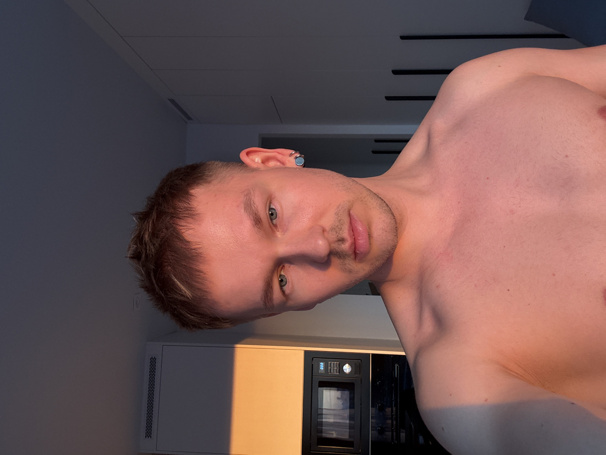 A shirtless man with short brown hair and a light beard looks at the camera. He has an earring in his left ear. The background shows a kitchen area with light-colored cabinets and a black microwave. The image may be taken in an indoor setting, with possible sunlight exposure. The location is unknown, including the possibility of Moscow, Russia.