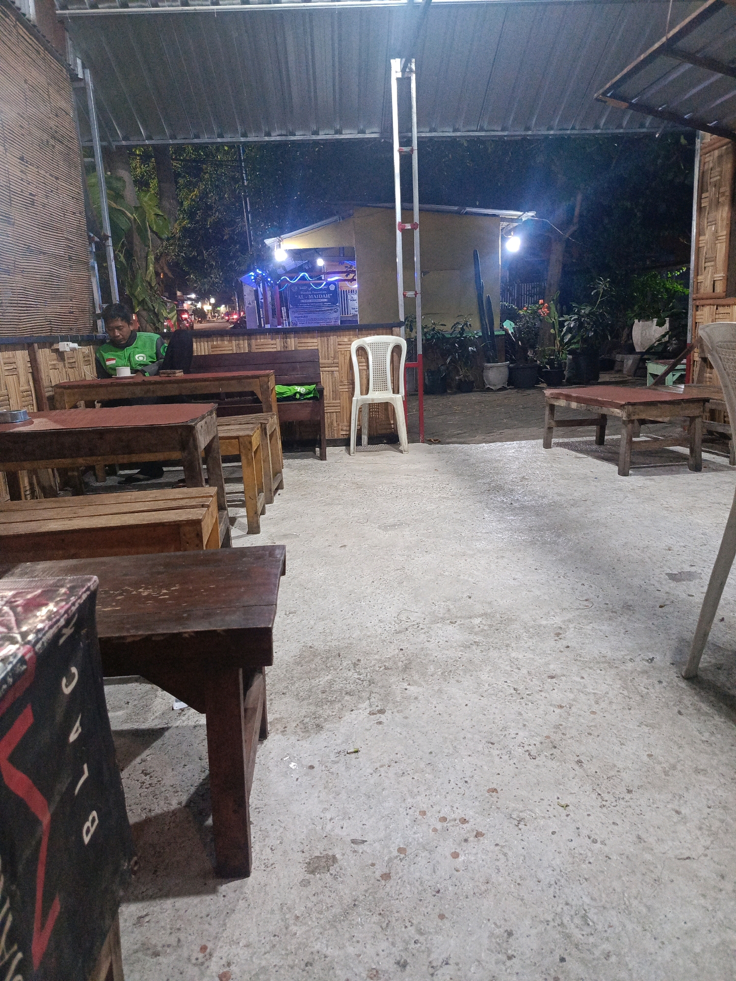 The image depicts an outdoor seating area of a cafe at night, possibly in Magetan, Indonesia, given the context. The area is covered by a corrugated metal roof, supported by metal poles. The walls of the cafe appear to be constructed from woven bamboo.
In the foreground, several wooden tables and benches are arranged on a concrete floor. One table in the lower left corner has a patterned cloth draped over it. Further back, a man wearing a green Gojek jacket is seated at a table, looking down at a cup in front of him. A white plastic chair is positioned near a table in the middle ground.
The background shows more seating and vegetation, with plants in pots. A building with yellow walls and blue decorative lights is visible behind the cafe. The lighting in the scene is dim, with some artificial lights casting shadows. The overall impression is that of a casual, open-air dining or resting space during the evening hours.