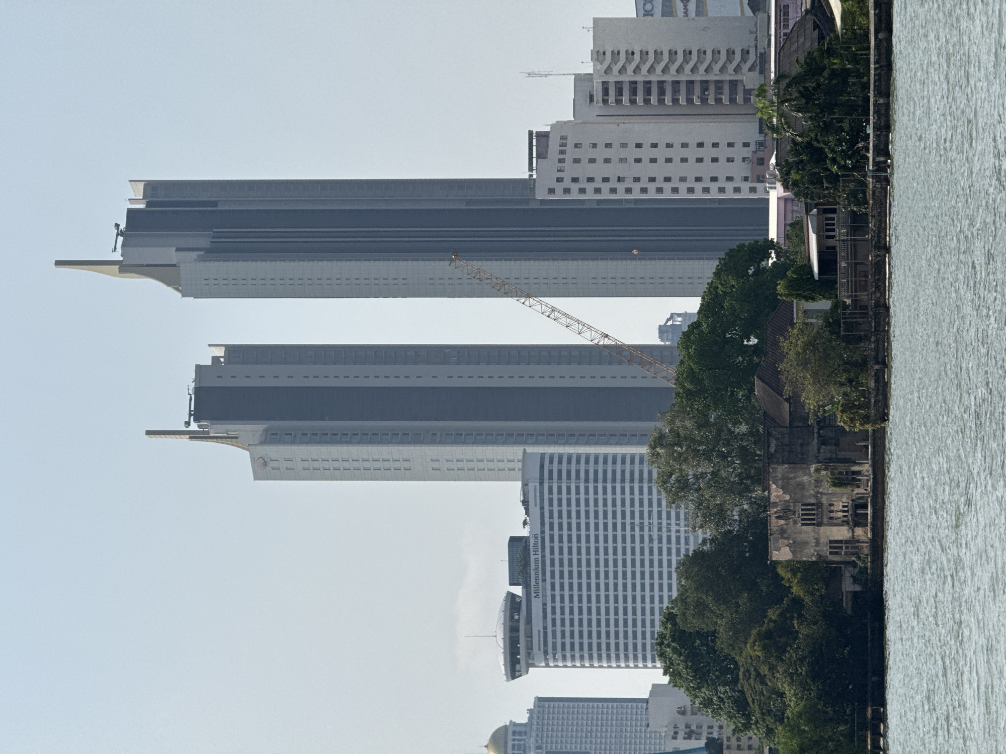 The image depicts a panoramic view of the Bangkok cityscape from the Chao Phraya River. In the foreground, choppy water occupies the lower quarter of the frame. The midground features a riverbank densely covered with green foliage and trees. Prominently situated on the bank is a dilapidated, multi-story concrete building with exposed, textured walls and multiple empty window apertures. To its right, partially obscured by trees, are smaller residential or commercial structures with various rooflines.
The background is dominated by modern high-rise buildings. Two towering, slender skyscrapers, identified as Magnolias Waterfront Residences at IconSiam, are centrally positioned, featuring grey facades and distinctive golden or bronze architectural accents at their apexes. A large, white "Millennium Hilton" hotel building with a prominent circular rooftop structure is visible to the left of these skyscrapers. An orange lattice boom construction crane extends diagonally upwards between the Millennium Hilton and the rightmost IconSiam tower. Additional multi-story buildings, including one with a golden dome on the far left, populate the distant skyline under a clear, pale sky. No people or discernible actions are visible in the scene.