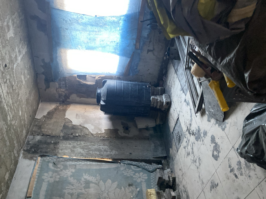 A dimly lit interior space reveals extensive disrepair. Plaster is crumbling from the walls, exposing the underlying structure. A black, cylindrical tank is precariously positioned on bricks along a wall. Tiled flooring is stained and cracked. Debris litters the space, including wood fragments and building materials. Natural light filters through a blue-toned opening above, casting shadows across the scene. The overall impression is one of neglect and decay.