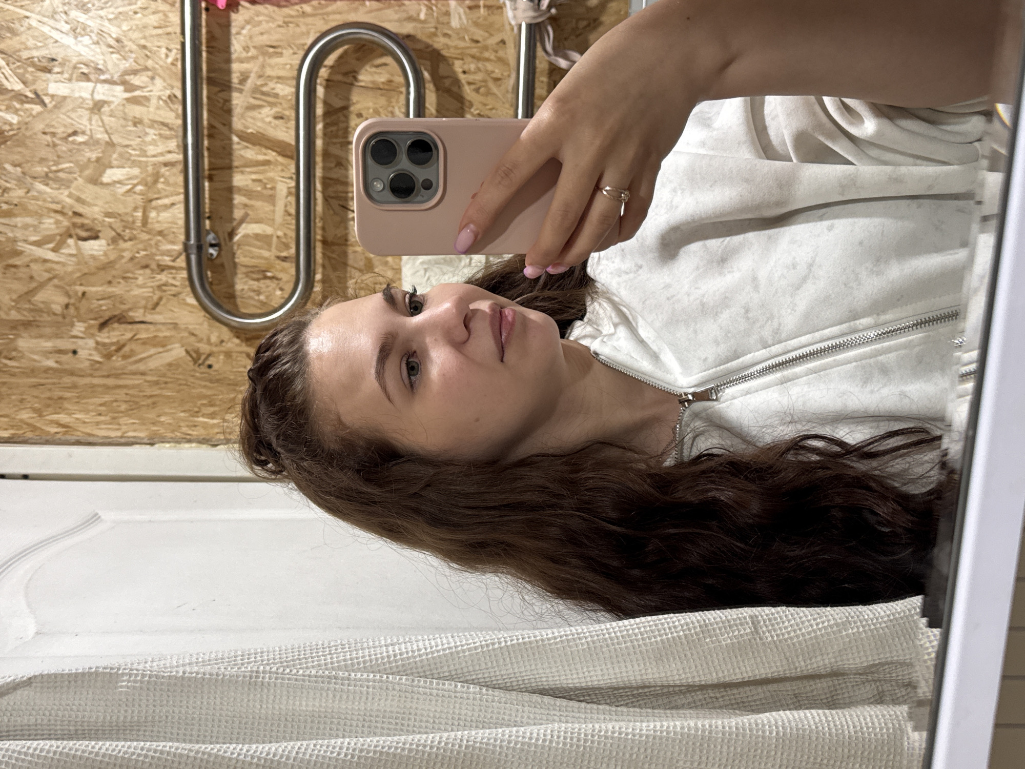 A woman is taking a selfie in a bathroom. The background includes a wooden wall, a towel rack, a white door, and a white textured towel. The location in the city of Nizhnyaya Gar', Russia is not verifiable.