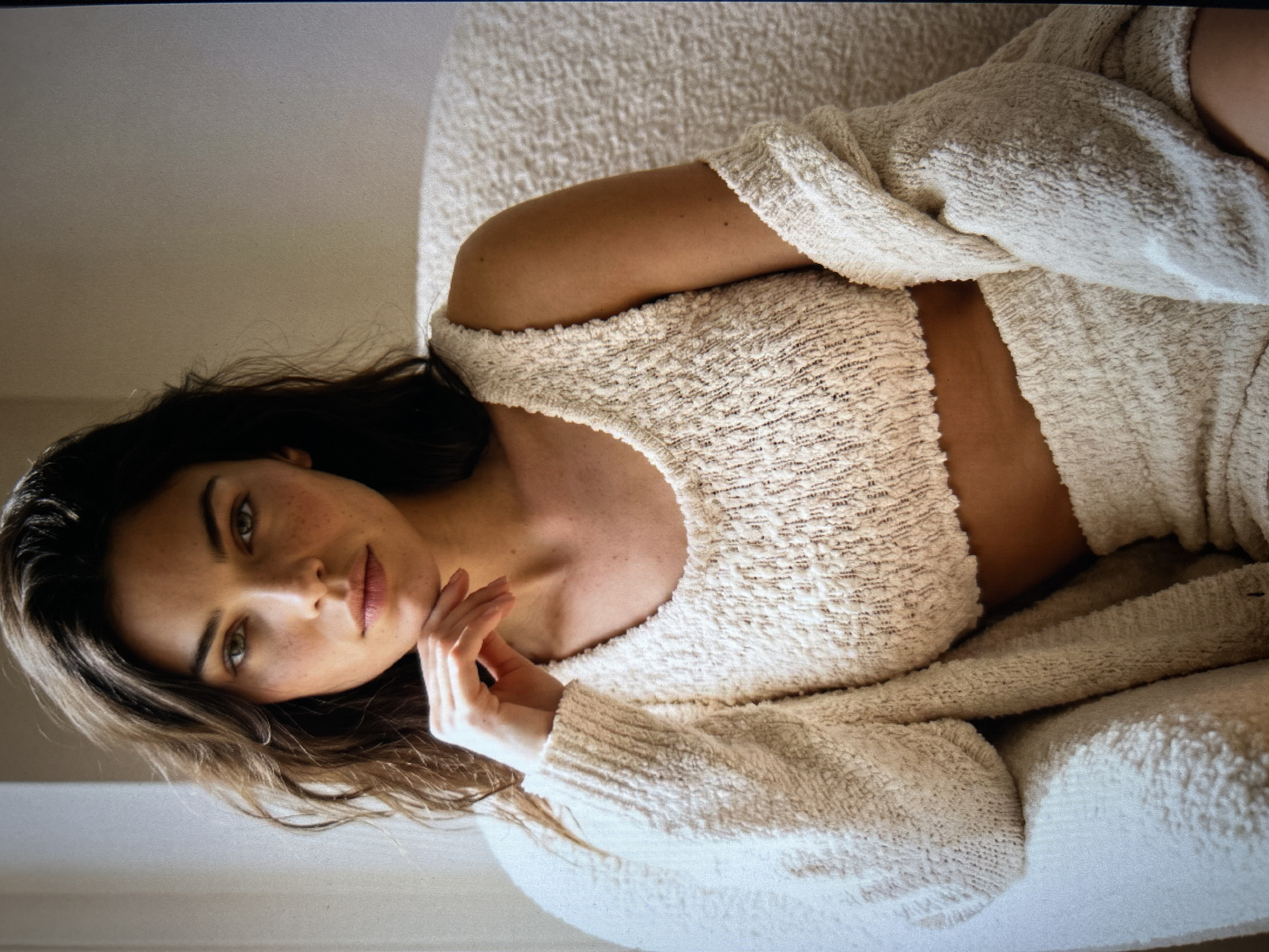 A medium shot shows a young woman with long, wavy brown hair sitting in a chair and looking at the camera. She is wearing a cream-colored, textured knit outfit consisting of a cropped tank top and a cardigan. Her right hand is resting on her chin, and her left arm is bent and resting on her lap. The background is a plain, light-colored wall, and a window is visible to the left, letting in soft light. The woman has freckles on her face and green eyes. The setting appears to be an indoor residential environment.