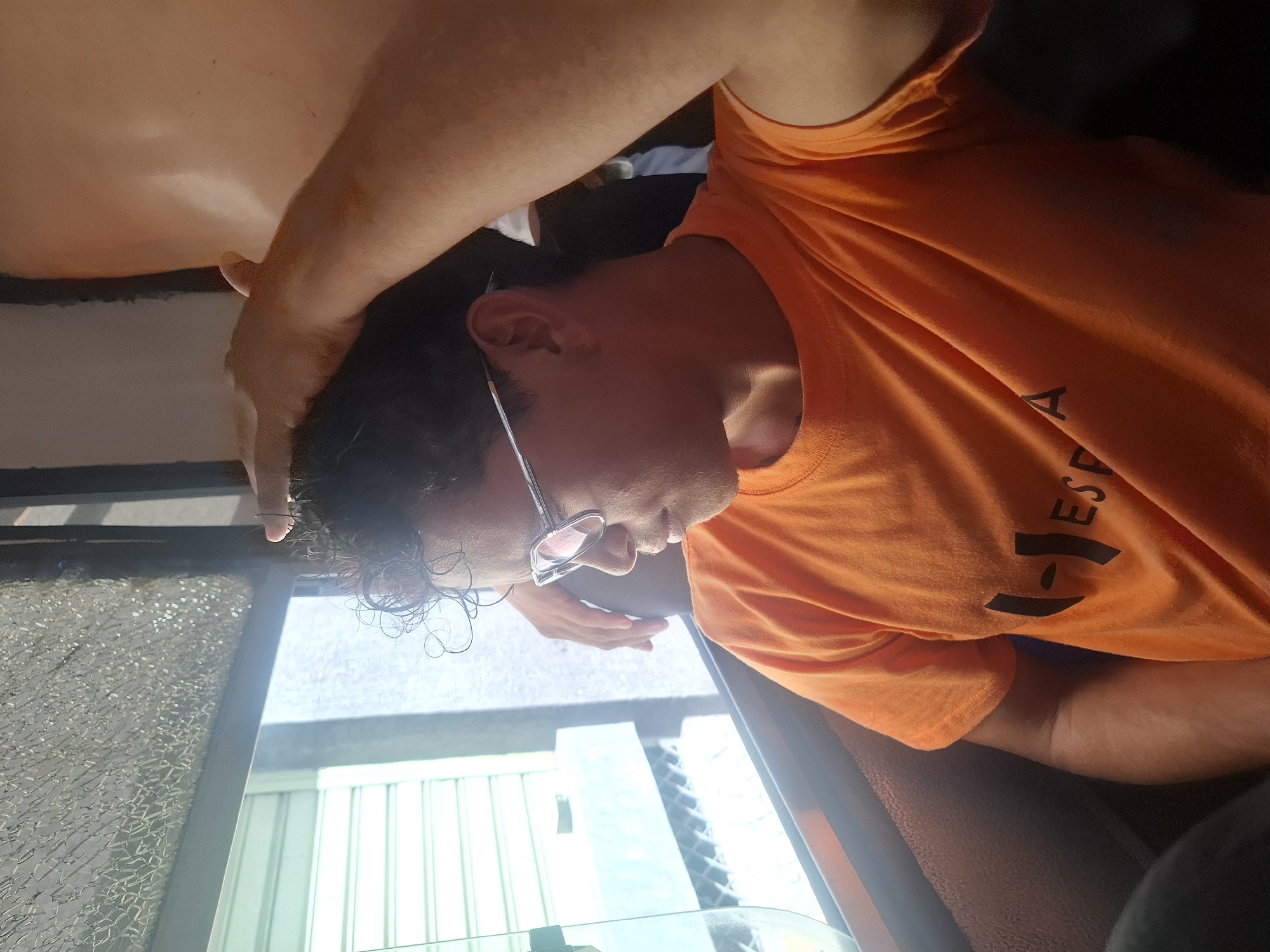 A man wearing an orange t-shirt and glasses is inside a vehicle with a cracked windshield. Outside the vehicle, there's a view of a building with vertical slats. The location is Juan Griego, Venezuela. The man has his hand on his head.
