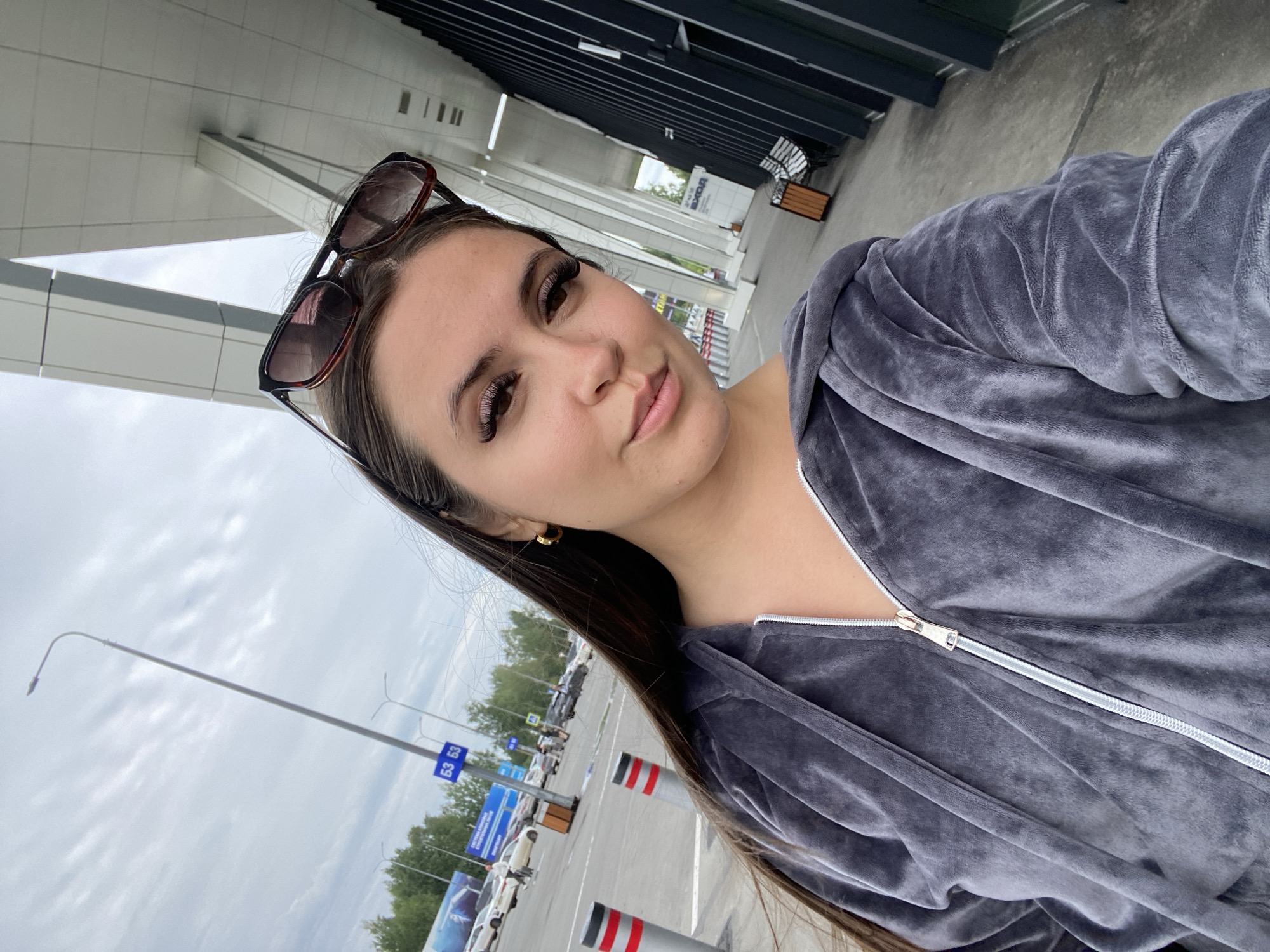 The image captures a young woman in a gray velour tracksuit, posing for a selfie outdoors. She has long dark hair and is wearing large sunglasses on top of her head. Her makeup is visible, including false eyelashes. In the background, a modern building with a black facade and white accents is visible. To the left of the building, there is a parking lot with several cars and a street lamp with blue signs displaying "53 53". The overall setting appears to be an urban or semi-urban environment, possibly near an airport or a large public facility. While the image has been associated with Новое Мартьяново, Russia, there are no specific landmarks or signs within the frame that definitively confirm this location. The lighting suggests it is daytime, with an overcast sky.