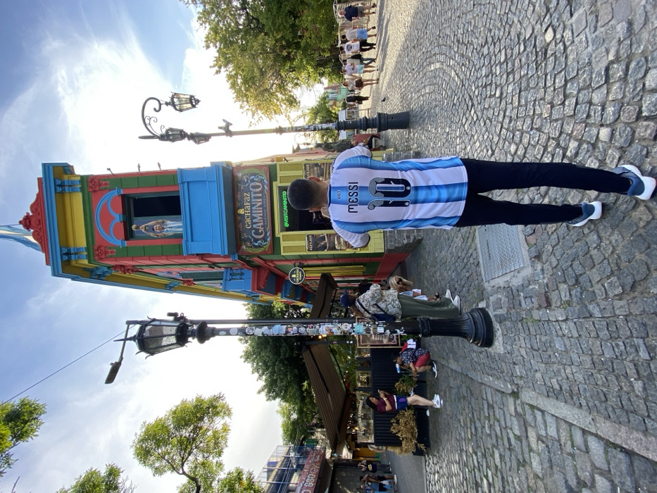 A tourist in a Messi jersey stands by a lamp post in La Boca, Buenos Aires, capturing a photo of the vibrant El Caminito building. The colorful structure features a statue in an upper window. Cobblestone streets lead to a distant group of people walking on the sidewalk. The day is bright, with a clear sky and trees providing partial shade to the bustling tourist destination.