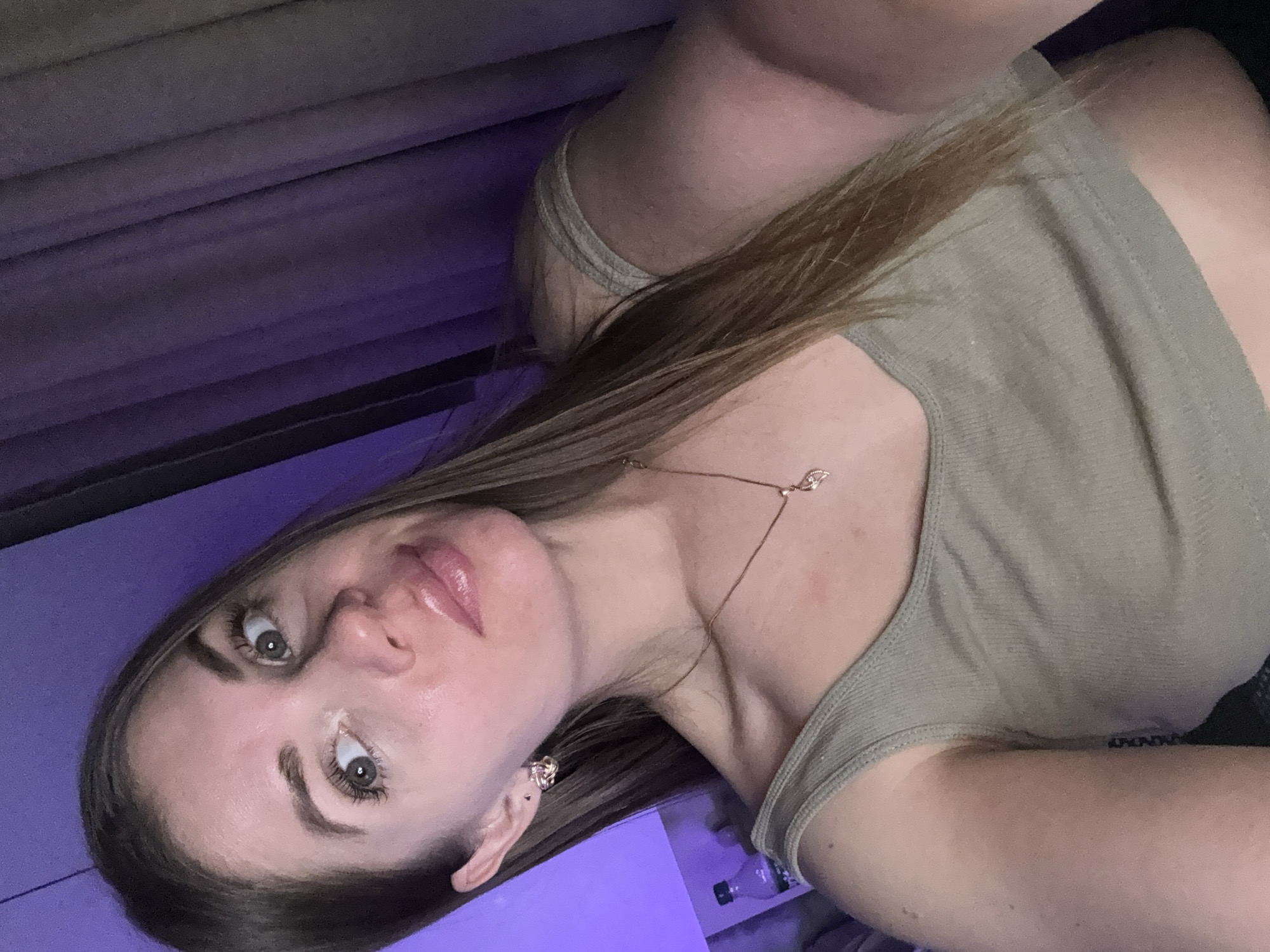A woman with long brown hair is seen in a selfie. She is wearing a light green tank top and a delicate gold necklace. Her makeup is subtle, with glossy lips. An earring is visible in her left ear. The background appears to be a room interior with gray walls and a purple light source. A water bottle is visible on a white surface. There is no information in the image that indicates the location as Batumi, Georgia.