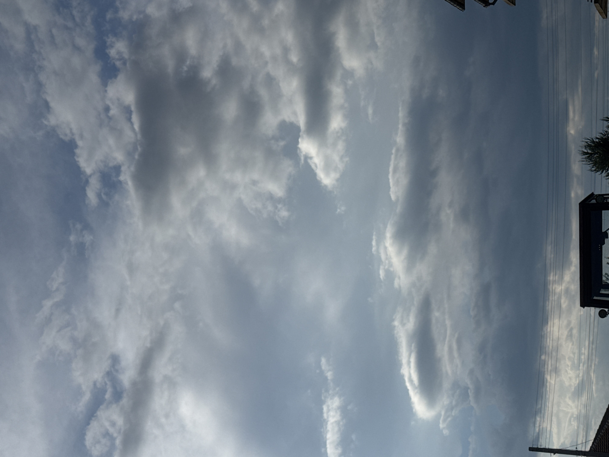 The image presents a view of a cloudy sky, presumably above the city of Sarnevo, Bulgaria, though there are no specific landmarks in the sky to confirm the precise location. The predominant feature of the image is the sky filled with various cloud formations, ranging from thin, wispy clouds to more substantial, gray masses. The clouds suggest potentially unsettled weather conditions. Power lines and parts of buildings are visible at the right side. The image is oriented vertically, and the details of the landscape, beyond the partial buildings and lines, are not visible.
