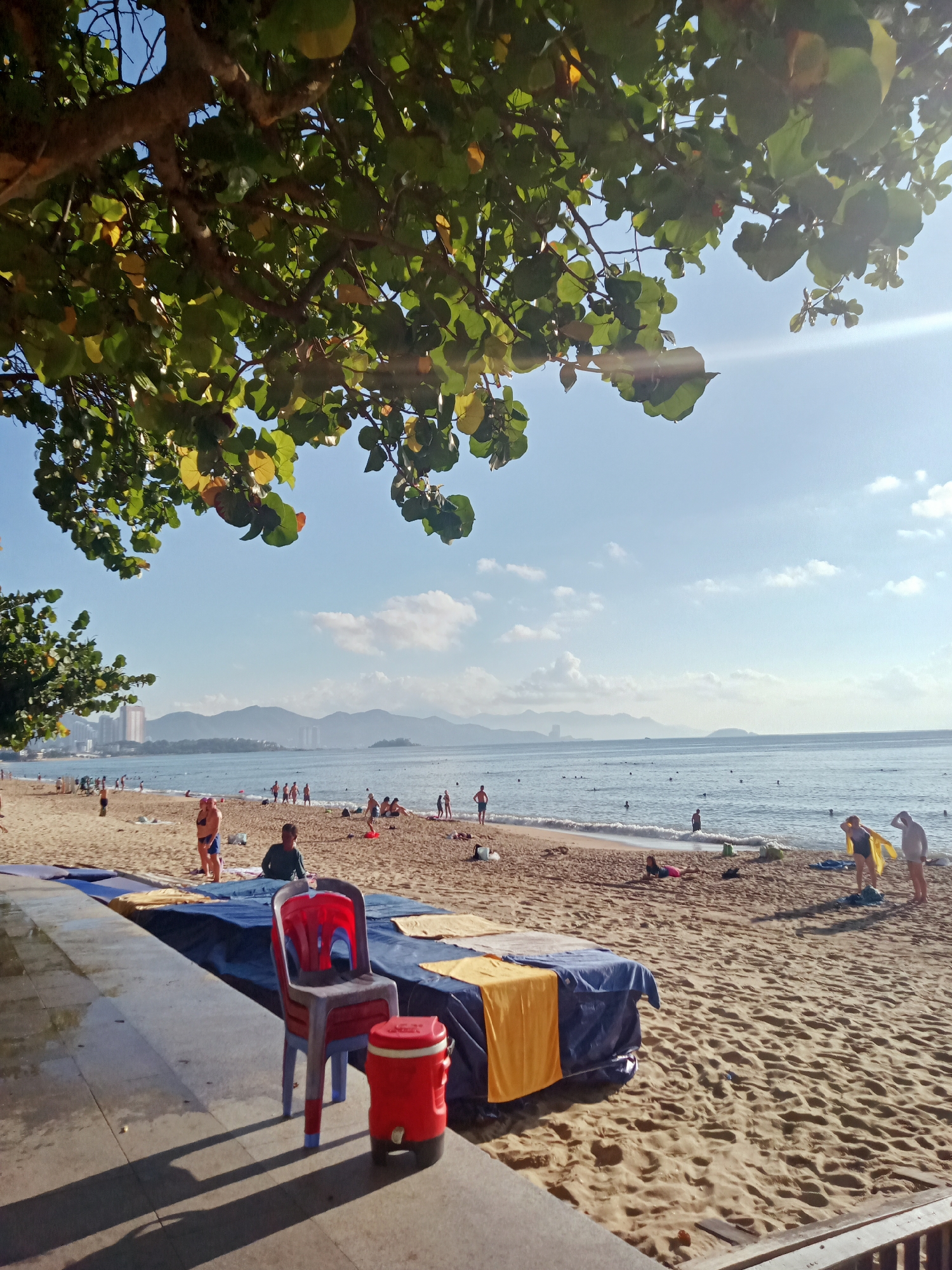 The image captures a sunny beach scene in Nha Trang Ward, Vietnam. In the foreground, a red plastic chair stands next to a red cooler on a concrete platform. Behind the platform, a beach scene unfolds with a blue tarp-covered surface draped with yellow towels. People are scattered across the sandy beach, some sunbathing, walking along the shore, or swimming in the ocean. In the background, the city skyline and mountains are visible under a clear blue sky with scattered clouds. Tree branches and leaves frame the upper portion of the image, partially obscuring the sky and creating dappled sunlight on the scene.
