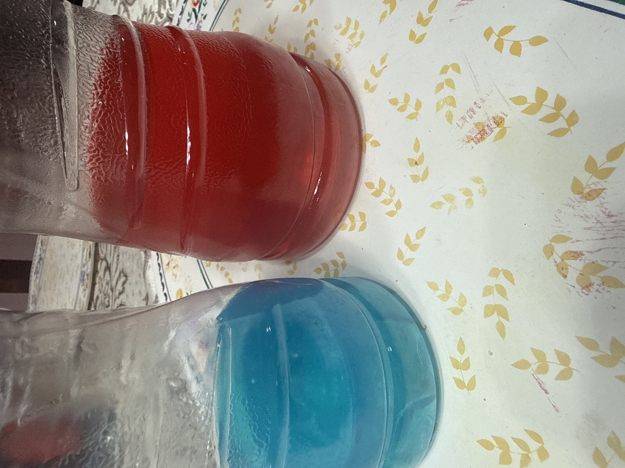 The image shows two glass bottles lying on their sides on a patterned surface. One bottle is filled with a red liquid, and the other with a blue liquid. The patterned surface appears to be a tablecloth or table covering with a white background and a repeating leaf design in yellow. The bottles are positioned next to each other, suggesting they might be related or part of the same setting. The image was taken in San Cristóbal, Mexico.