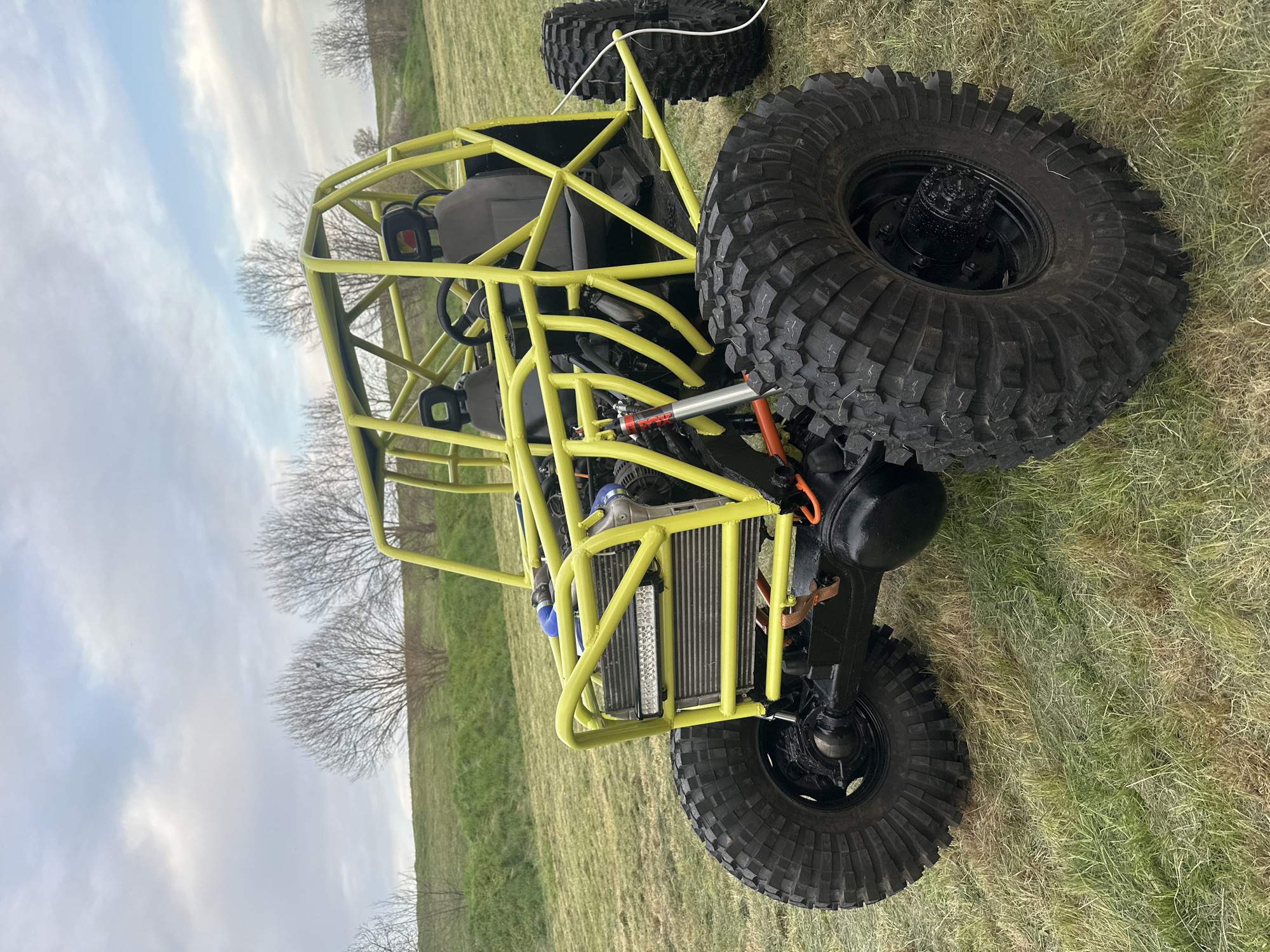 A yellow tubular frame all-terrain vehicle sits on a grassy field in an indeterminate location that is not identifiable as Choba, Bulgaria. The vehicle has oversized black tires, a front-mounted radiator, and two bucket seats. The background includes a horizon line of bare trees against a cloudy sky.
